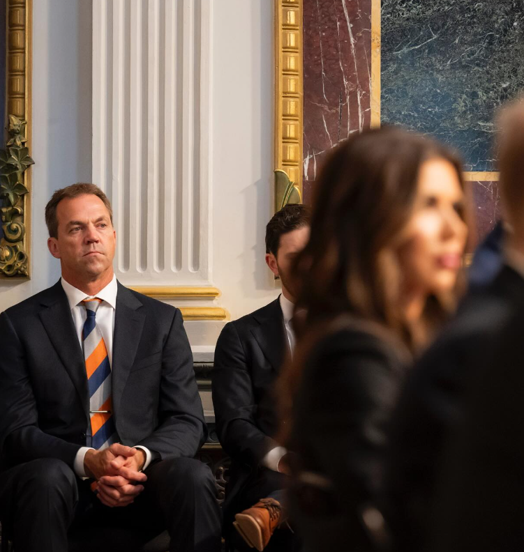 Bryon Noem sits with his hands clasped inside a formal interior setting, appearing focused as others are seated nearby. | Source: Instagram/kristinoem