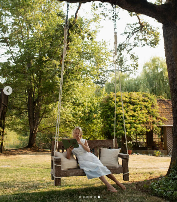 Pamela Anderson reclines barefoot on a suspended wooden porch swing under the dappled shade of a sprawling tree at Arcady, lost in a book with cozy outdoor pillows beside her. Celebrating her plant-based series "Pamela's Cooking with Love," she shared the peaceful farm moment with the caption, "Cooking with Love… our little show that’s everything I could ask for—my garden, a little farm on Vancouver Island, and chefs from all over the world. 👩🌾… It’s been a dream come true." | Source: Instagram/pamelaanderson