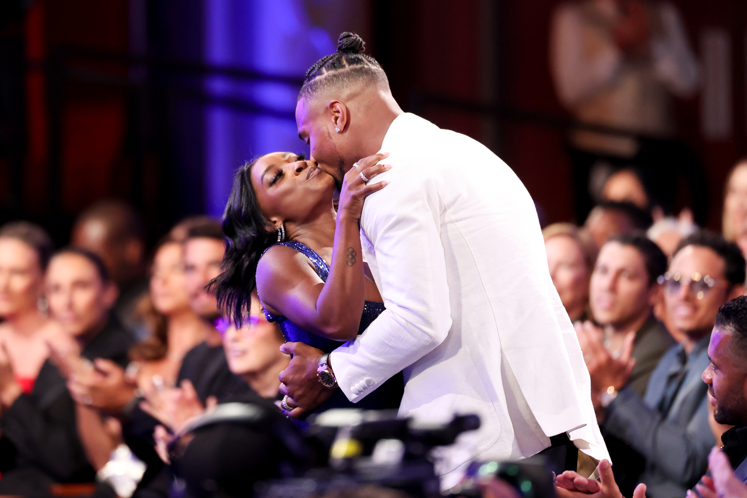 Jonathan Owens kisses Simone Biles as she stands to accept an award, surrounded by a cheering audience. Biles leans into the embrace with one arm around him, while attendees applaud the moment, capturing a mix of celebration and affection inside the venue.