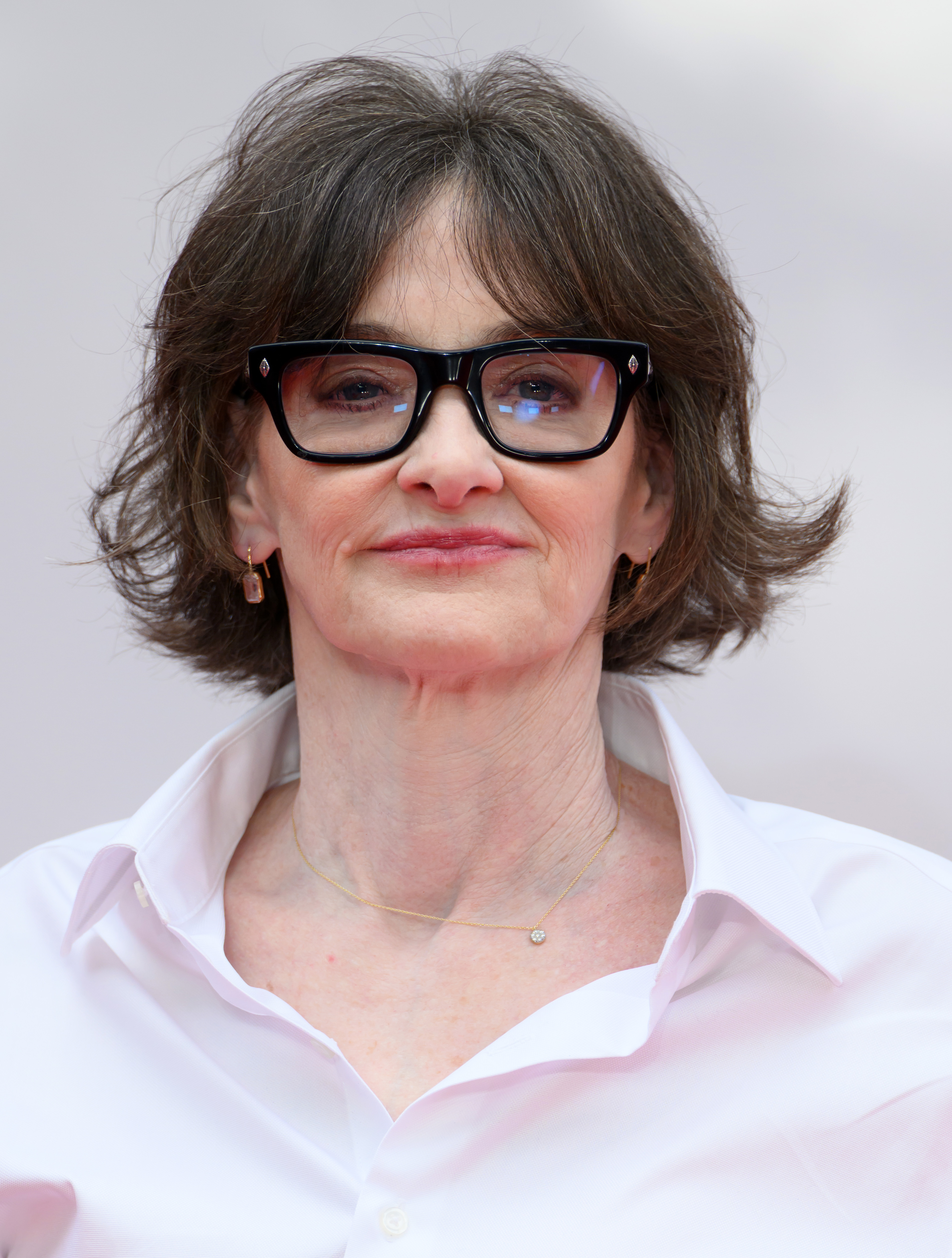 Joan Cusack is captured in a close-up portrait, her signature glasses framing her face against a soft neutral backdrop. The tight crop draws attention to her calm expression, tousled hairstyle, and understated jewelry.