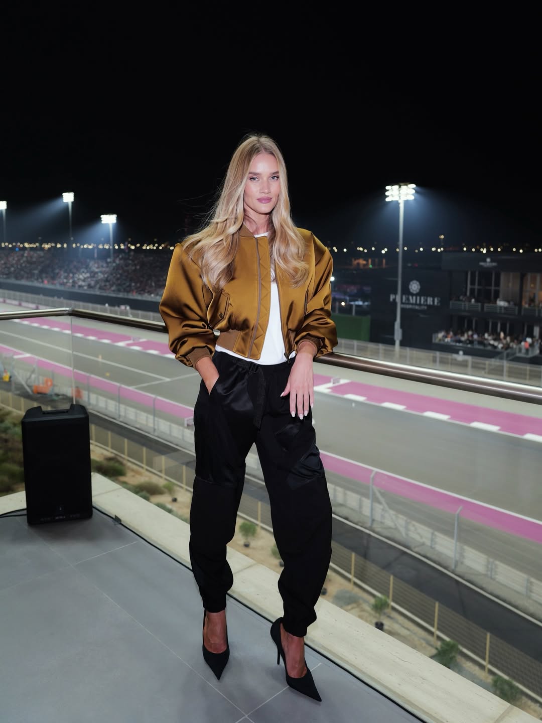 Rosie Huntington-Whiteley poses on a balcony overlooking a brightly lit racetrack at night, blending sporty glamor with effortless polish. | Source: Instagram/rosiehw