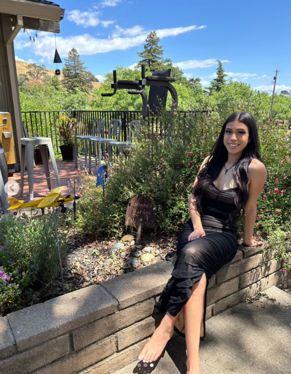 Marissa DiNapoli sits outside in a garden area, smiling in a black dress near a sign that reads "Karlene's Garden," posted on July 1, 2025 | Source: Instagram/moniice__