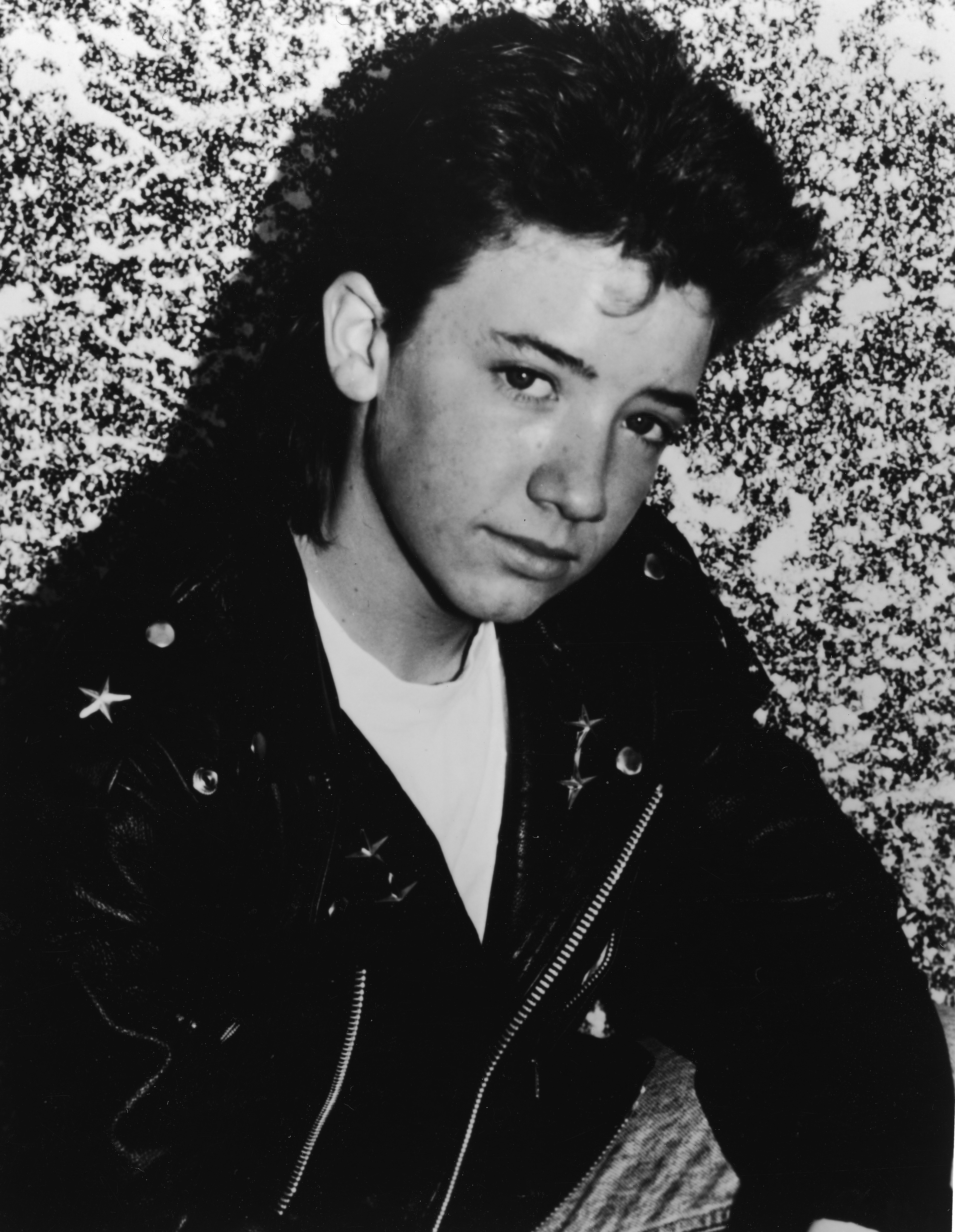 David Faustino channeled edgy retro style in a black-and-white promotional portrait for the television series "Married... with Children" circa 1990. Leaning forward with a cool, deadpan stare and his signature mullet hairstyle, the young actor sported a stud-embellished leather motorcycle jacket layered over a plain white crewneck tee. | Source: Getty Images