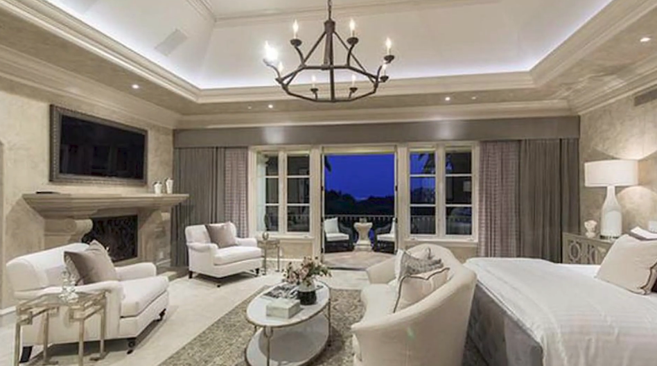 The primary suite centers on a carved stone fireplace with an integrated flat-screen television, a tray ceiling illuminated by ambient cove lighting and an iron chandelier, and French doors leading to a private balcony. | Source: YouTube/Taste of Country