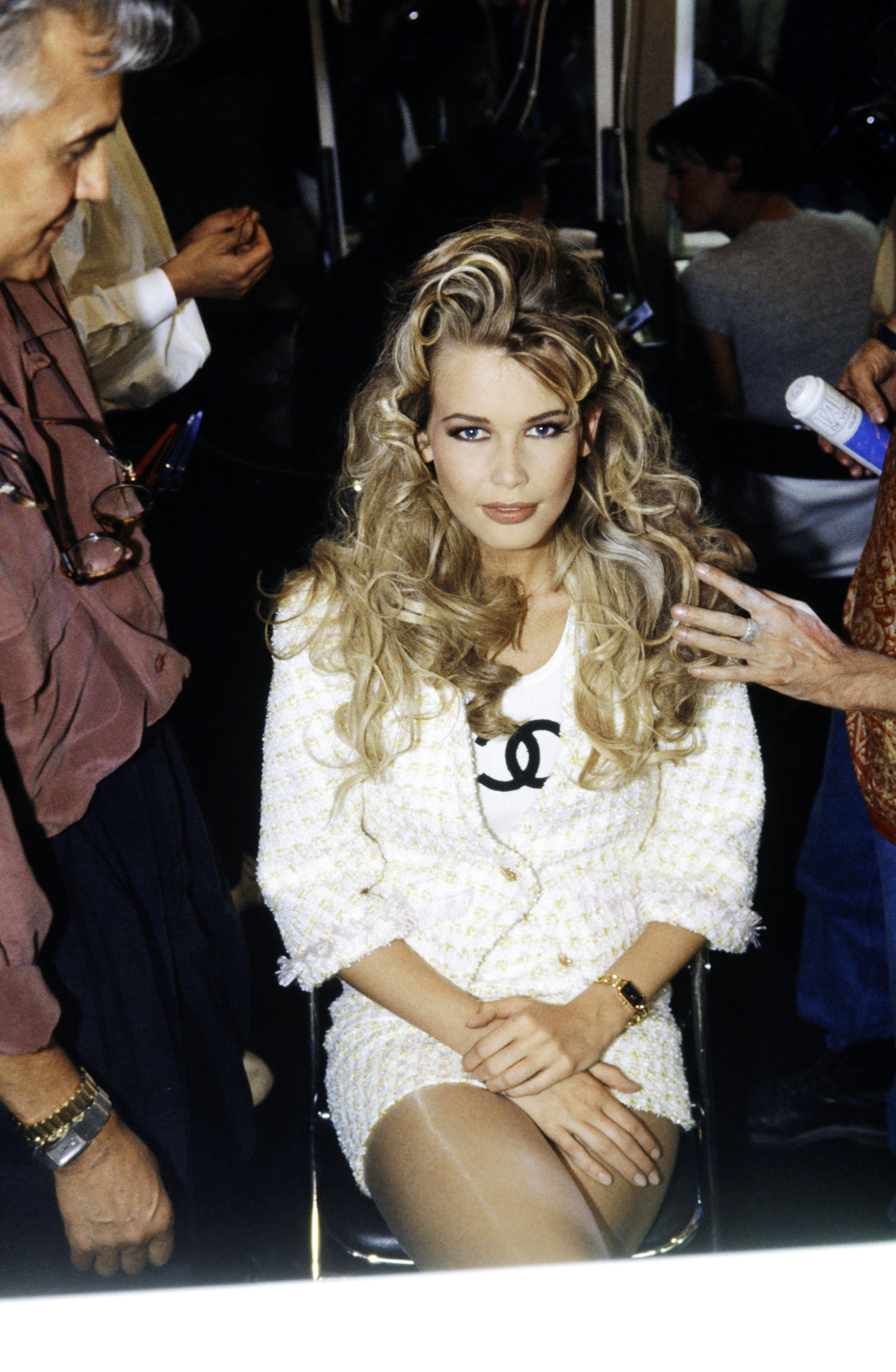 Backstage at Chanel's Spring Couture Show, Claudia Schiffer radiates poise in a chic tweed ensemble, her golden curls styled to perfection. Surrounded by the bustle of preparation, she embodies the timeless elegance that defined the era's fashion.