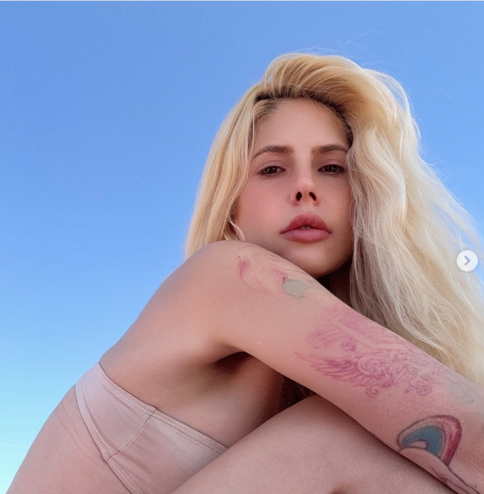 A sun-drenched outdoor portrait against a clear blue sky, shared to Instagram on August 15, 2025. Gazing directly into the camera, the musician wore a strapless nude top and showed off the colorful illustrative tattoos along her forearm, with voluminous platinum blonde hair swept gracefully in the breeze.