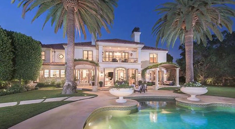 Towering palm trees frame the resort-style backyard at twilight, illuminating the glowing freeform swimming pool, shaded dining pergolas, and warm French doors along the mansion's illuminated rear facade. | Source: YouTube/Taste of Country