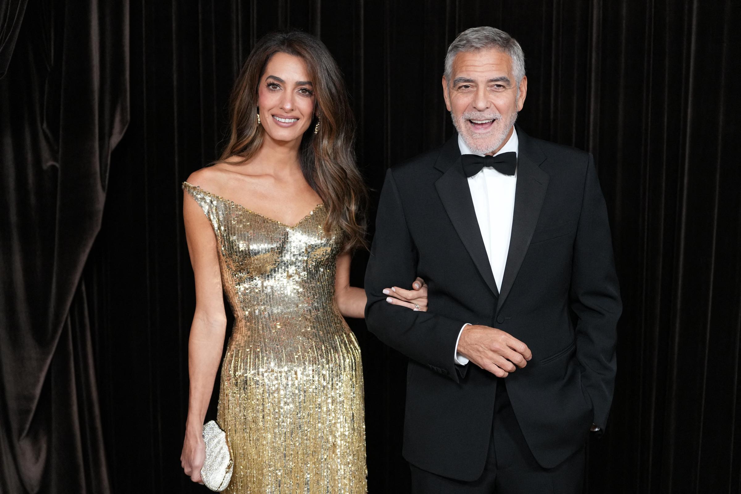 George and Amal Clooney pose arm in arm at the 2026 Albies in London on October 9. Amal dazzles in a shimmering gold, off-the-shoulder gown with a matching clutch, while George wears a classic black tuxedo with a bow tie. | Source: Getty Images