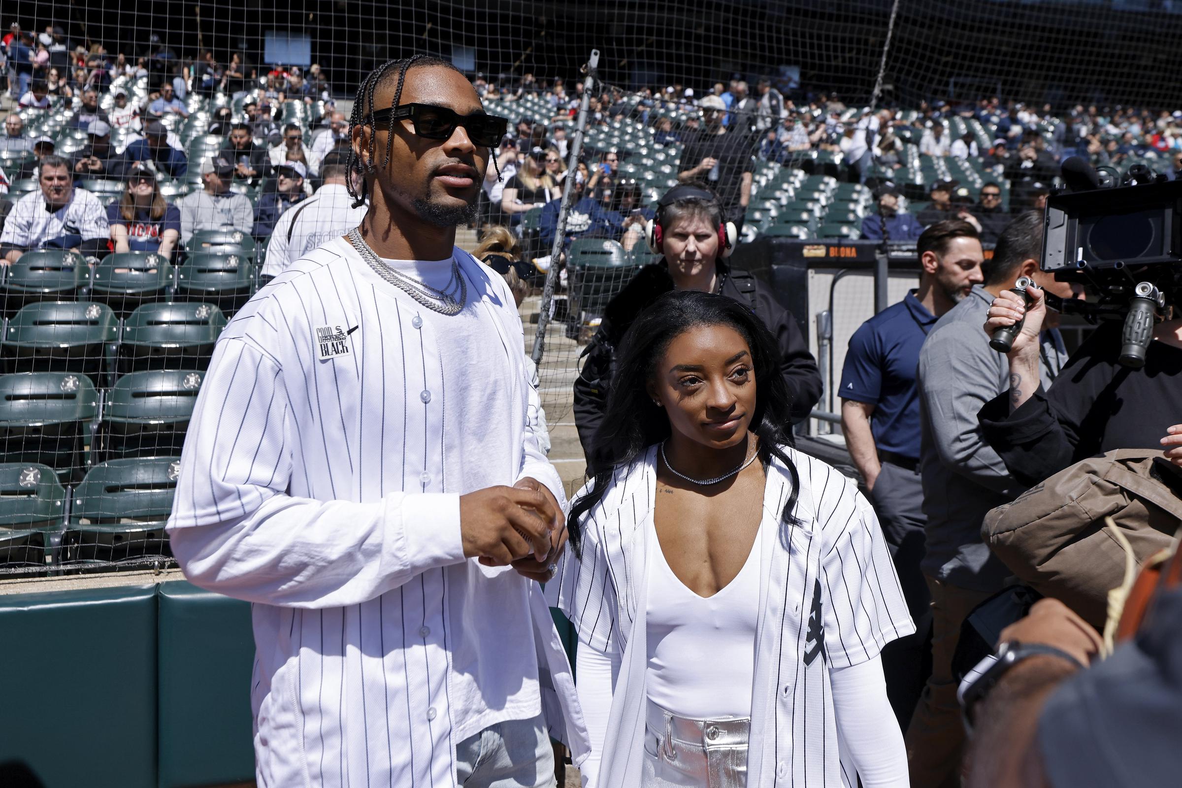 Jonathan Owens and Simone Biles are seen standing near the field ahead of the Chicago White Sox vs. Cincinnati Reds game at Guaranteed Rate Field on April 13, 2024. Both wear matching pinstripe jerseys, with Owens in sunglasses and layered chains, while Biles pairs hers with a white top and metallic silver pants.