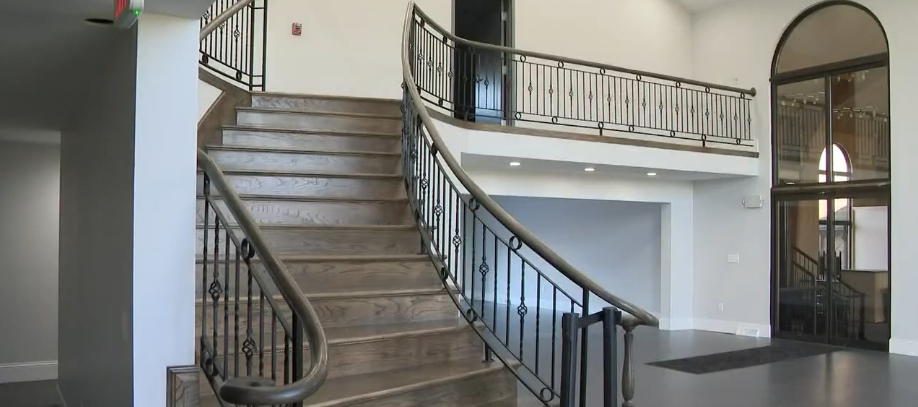 The mansion's sweeping staircase survived its transition from Mike Tyson's private home to Living Word Sanctuary Church, but the surrounding space received a substantial update. Restored wooden steps rose between dark metal railings toward the second-floor balcony, while crisp white walls and clean flooring replaced the worn finishes seen before renovation, preserving a prominent architectural feature within the property's completely reworked interior design. | Source: YouTube/WKYC Channel 3