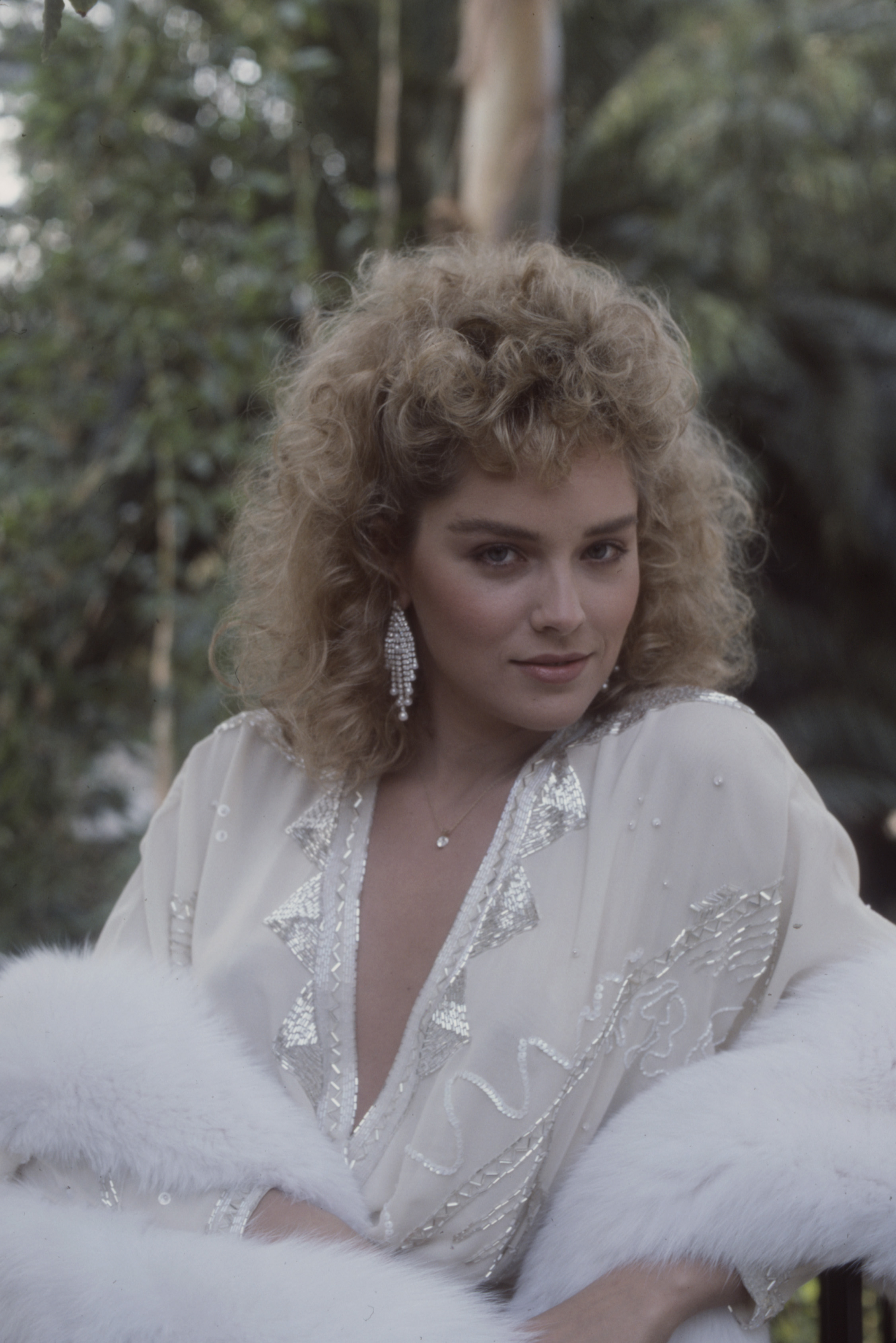 Sharon Stone gazes at the camera in a glamorous still from the ABC TV movie "Calendar Girl Murders." Wearing a white, silver-trimmed outfit and dramatic earrings, she appears surrounded by greenery.