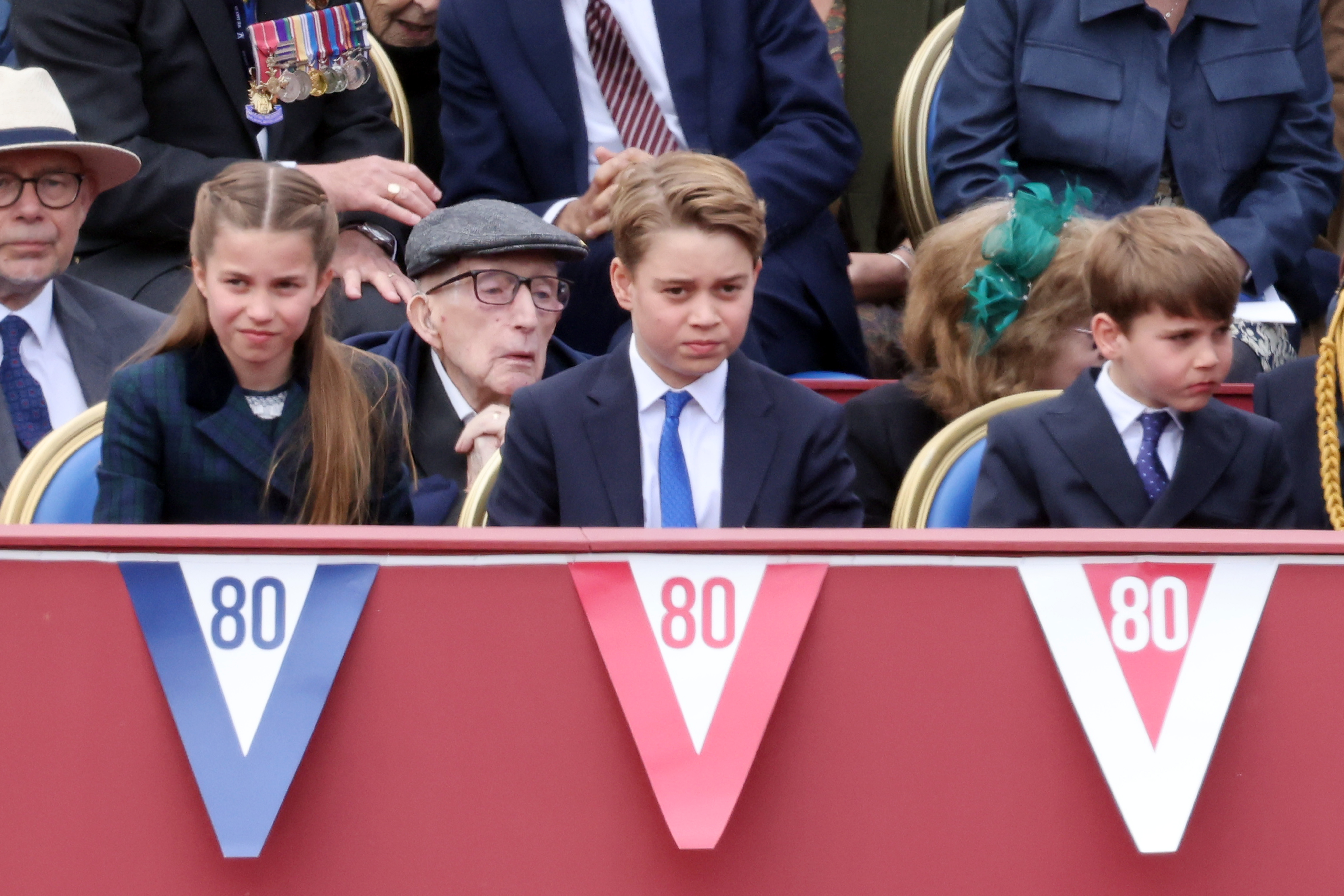 Princess Charlotte, Prince George, and Prince Louis sit side by side during the VE Day ceremony, each wearing a formal expression tinged with boredom. Their subtle frowns, fixed gazes, and slightly pursed lips reflect the long, solemn tone of the commemorative event.