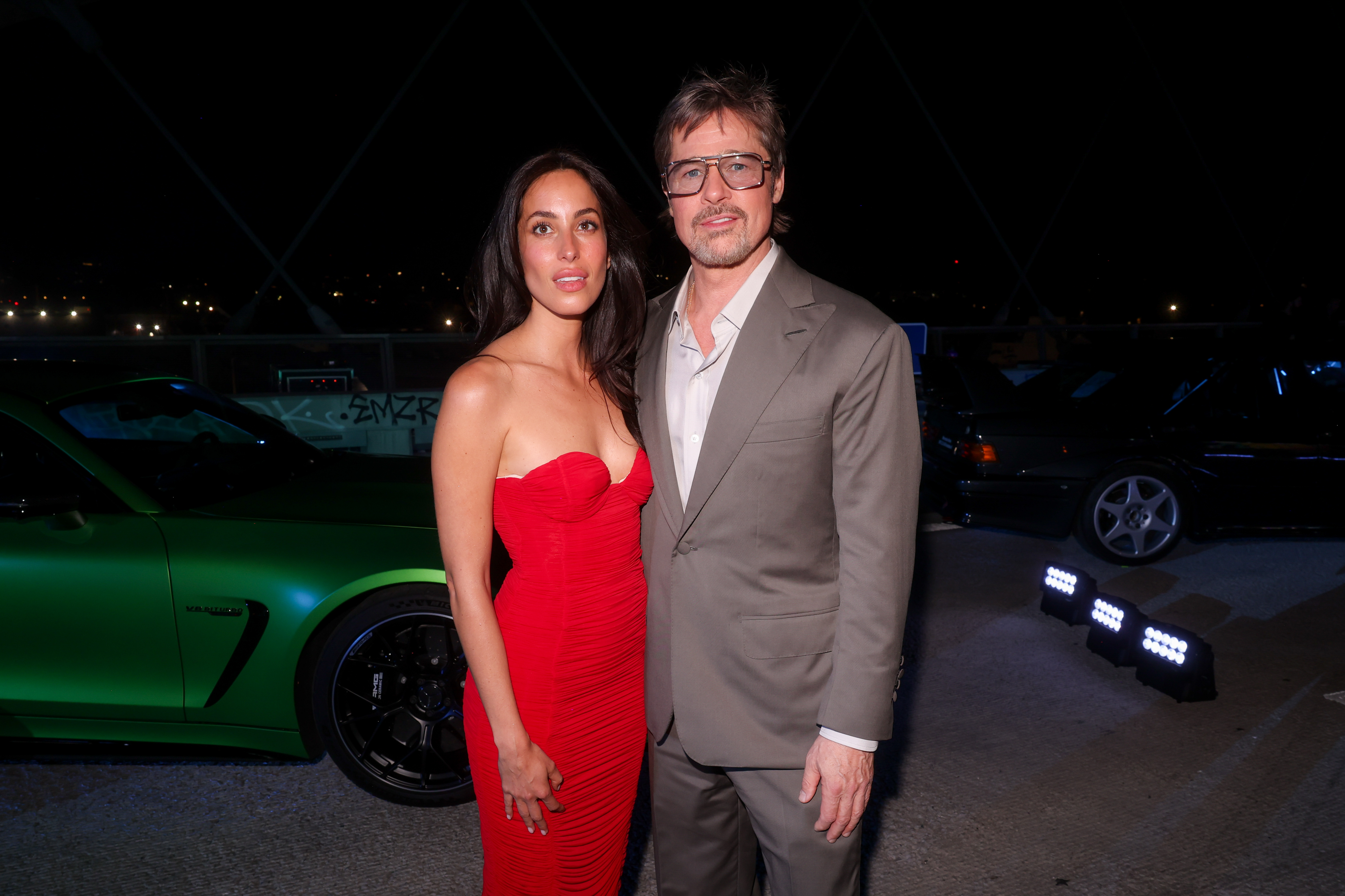 Ines de Ramon poses beside Brad Pitt in front of a green Mercedes-AMG vehicle, adding to the high-end racing atmosphere surrounding the glamorous premiere event.