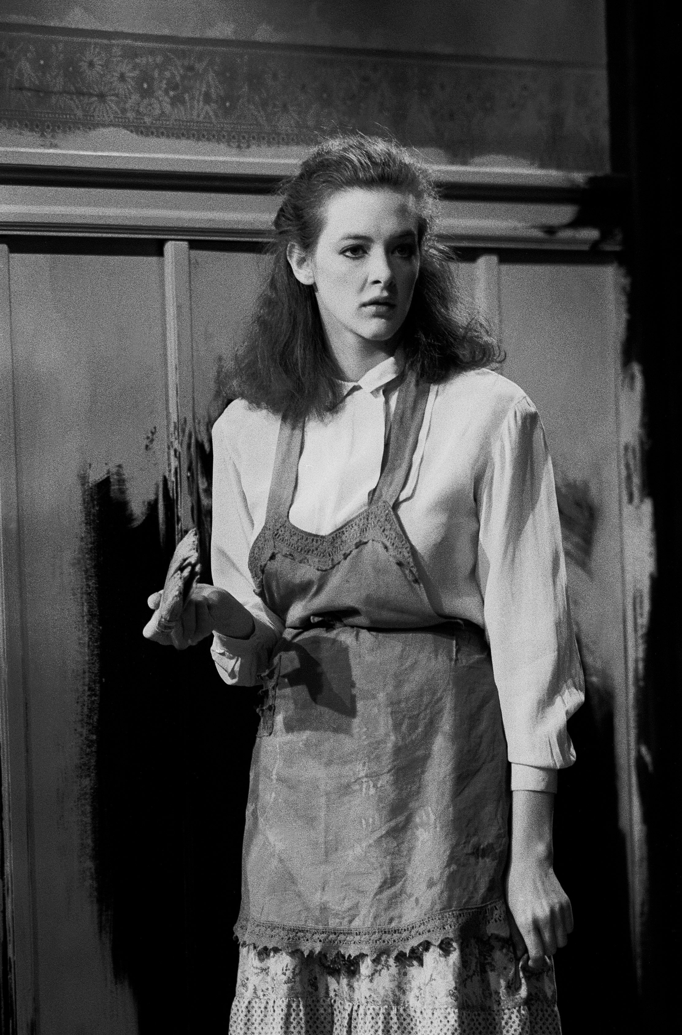 Joan Cusack appears deep in character, standing in a vintage-style outfit with an apron layered over a buttoned blouse. Her thoughtful expression and poised stance give the scene a quietly dramatic feel.