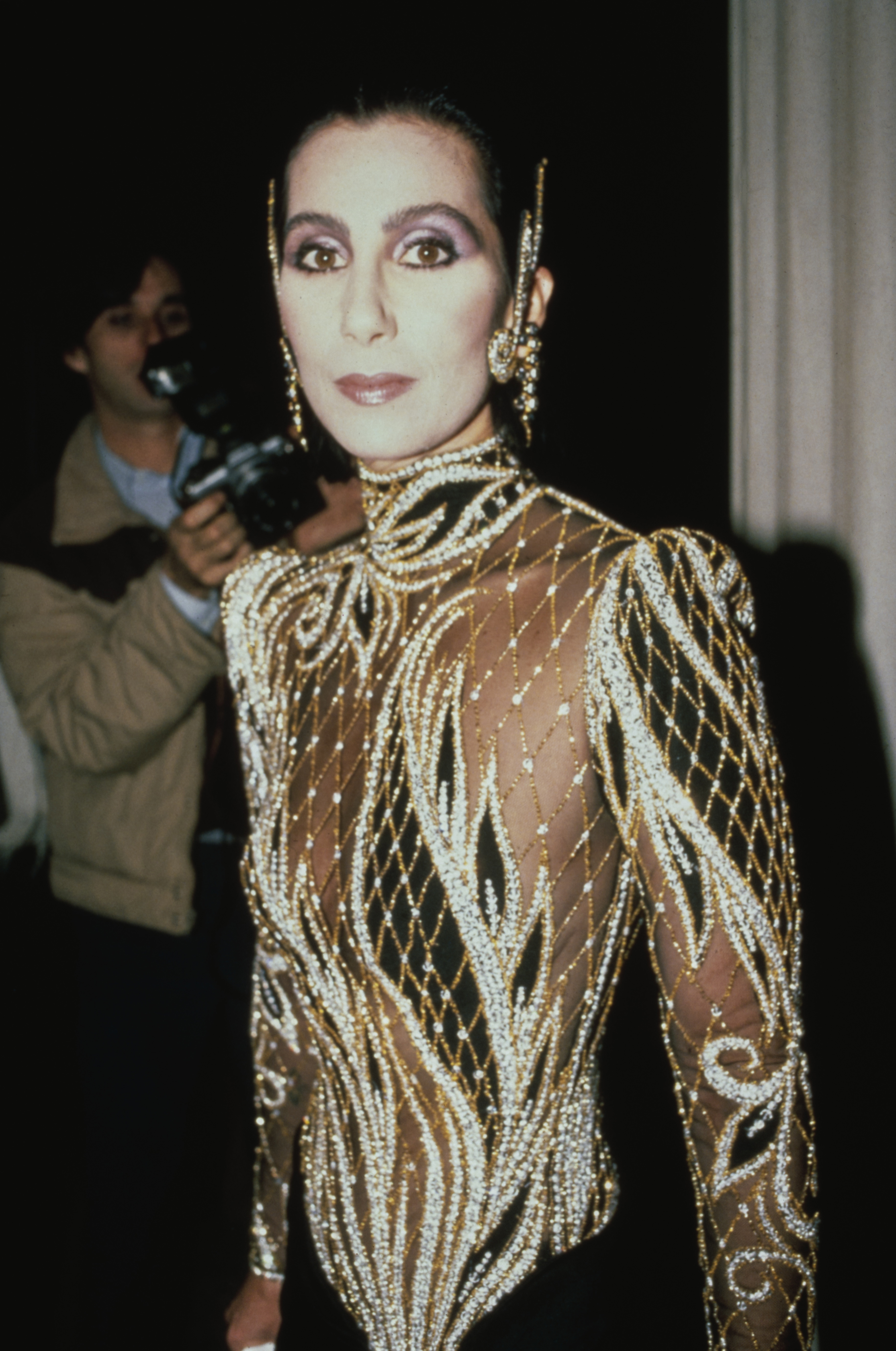 Cher made an unforgettable fashion statement in a custom beaded black-and-sheer creation by Bob Mackie at the Met Gala exhibition of "Costumes of Royal India" on December 9, 1985, in New York City. The high-neck design featured intricate, swirling gold and silver rhinestone lattice embroidery across a sheer bodice with padded shoulders, accented by striking ear-cuff jewelry and dramatic evening makeup. | Source: Getty Images