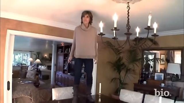 Lisa Rinna stands directly on top of her formal dining table beneath a wrought-iron chandelier. | Source: YouTube/@Biography