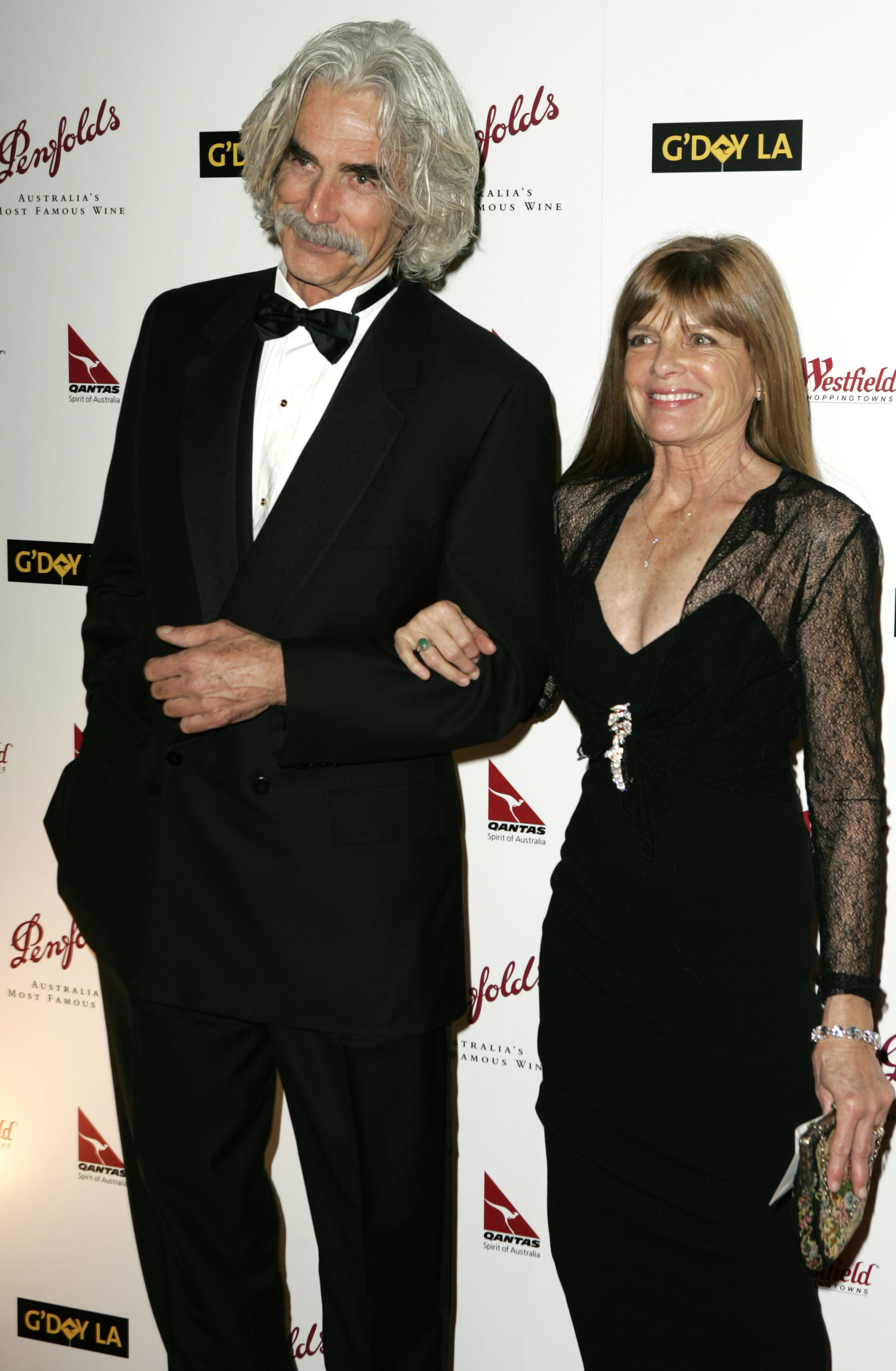 Sam Elliott and Katharine Ross coordinated in timeless black attire at the 2nd Annual Penfolds Gala Black Tie Dinner on January 15, 2005, hosted at the Century Plaza Hotel in Los Angeles, California. Katharine linked her arm through his while donning an elegant black gown with sheer lace long sleeves, a plunging neckline, and a sparkling crystal brooch at the bodice. Beside her, Sam stood sharp and distinguished in a double-breasted tuxedo and bow tie, sporting his trademark bushy gray mustache and wavy silver hair. | Source: Getty Images