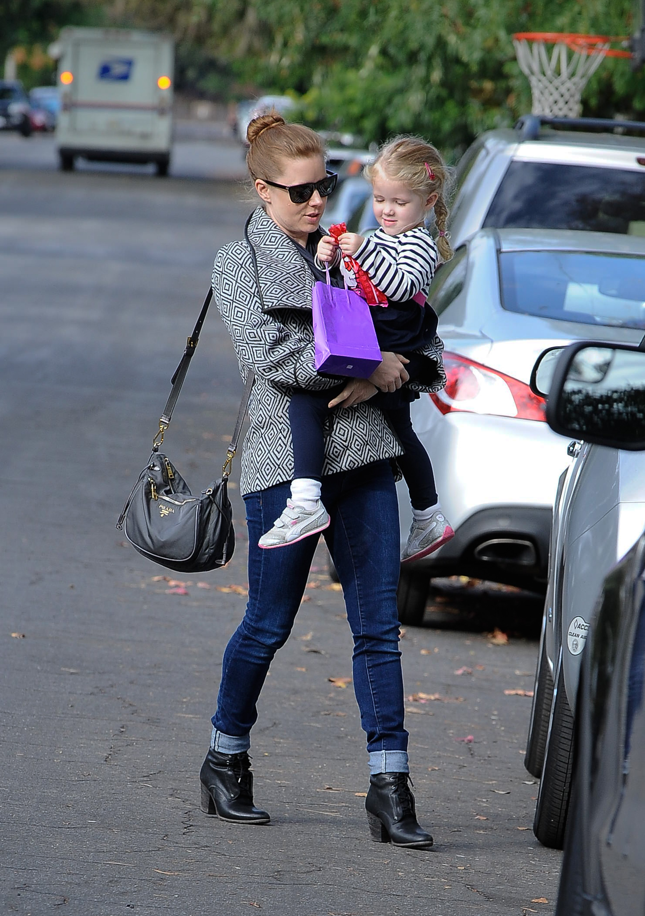 Amy Adams carries her daughter, Aviana Olea Le Gallo, while walking along a Los Angeles street. Dressed casually and wearing sunglasses, Amy balances a handbag as Aviana holds a small purple gift bag.