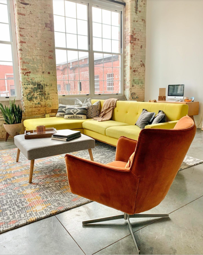 A more colorful take on the loft shows a mustard-yellow sectional and rust-toned chair set against exposed brick and oversized windows, with layered furnishings adding warmth to the industrial space. | Source: Facebook/Judson Mills Loft