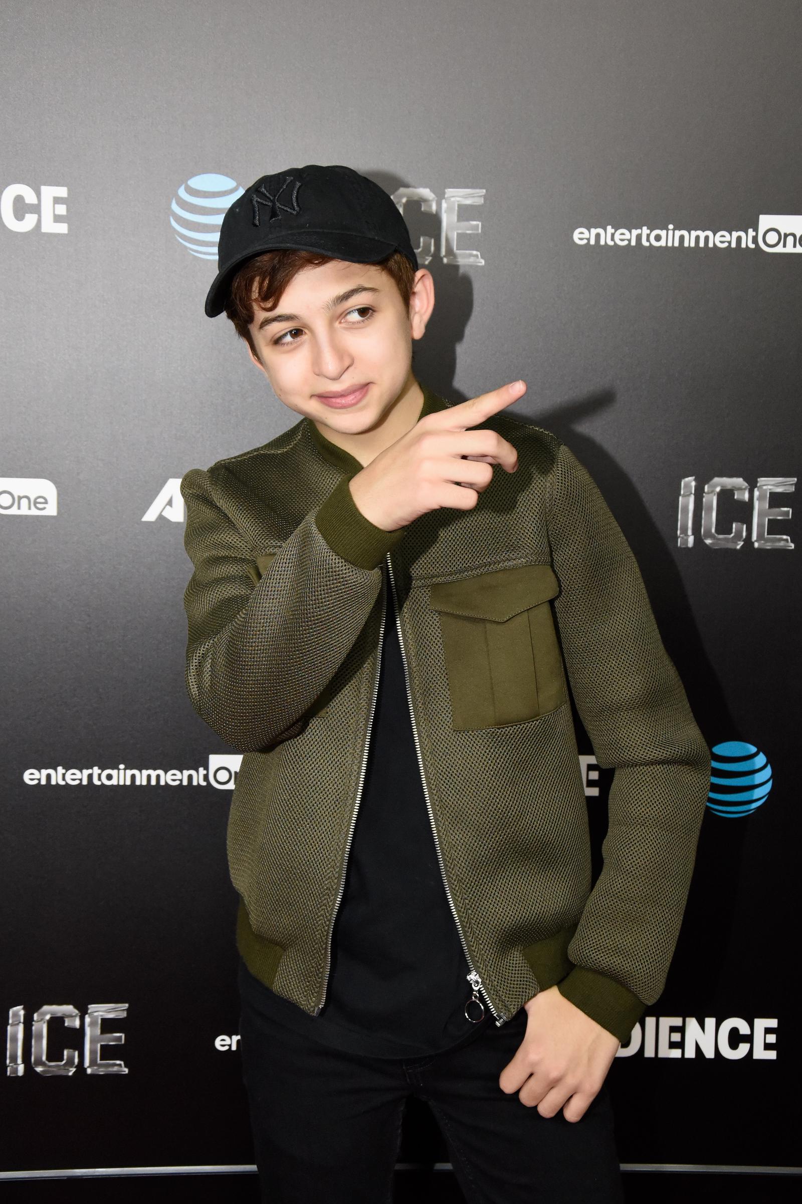 J.J. Totah arrives at the premiere of Audience Network's Ice at ArcLight Cinemas in Hollywood, California, wearing an olive-green textured jacket, a black baseball cap, and dark trousers. Totah points to the side while posing for photographers. | Source: Getty Images
