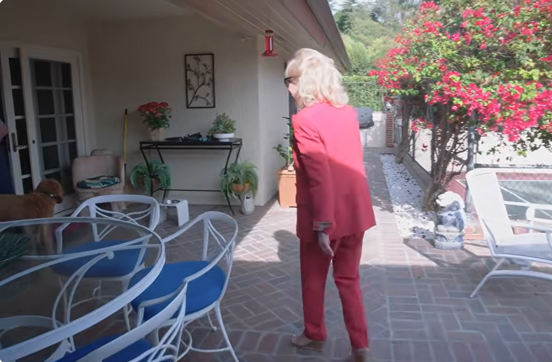 Barbara Eden walks across her covered patio toward the garden, wearing a pink-red pantsuit and sunglasses. White metal chairs with blue cushions, potted plants, and vibrant pink flowering shrubs surround the brick-paved outdoor seating area. | Source: YouTube/People
