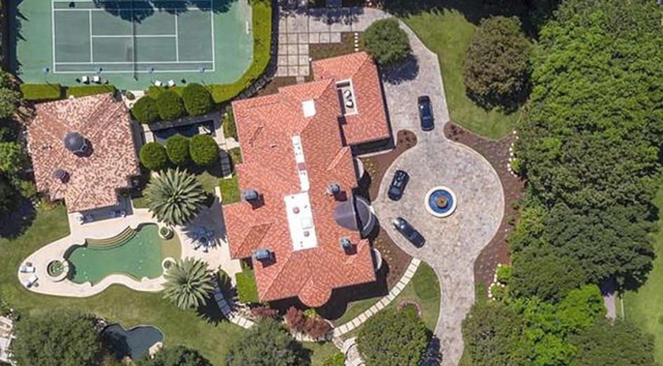 An aerial view highlights the expansive layout of Reba McEntire's former 1.8-acre Beverly Park estate, showcasing the main residence's terracotta tile roofline alongside a circular cobblestone motor court, a lighted tennis court, a detached poolside pavilion, and a Grecian-style swimming pool surrounded by lush privacy hedging and mature trees. | Source: YouTube/Taste of Country