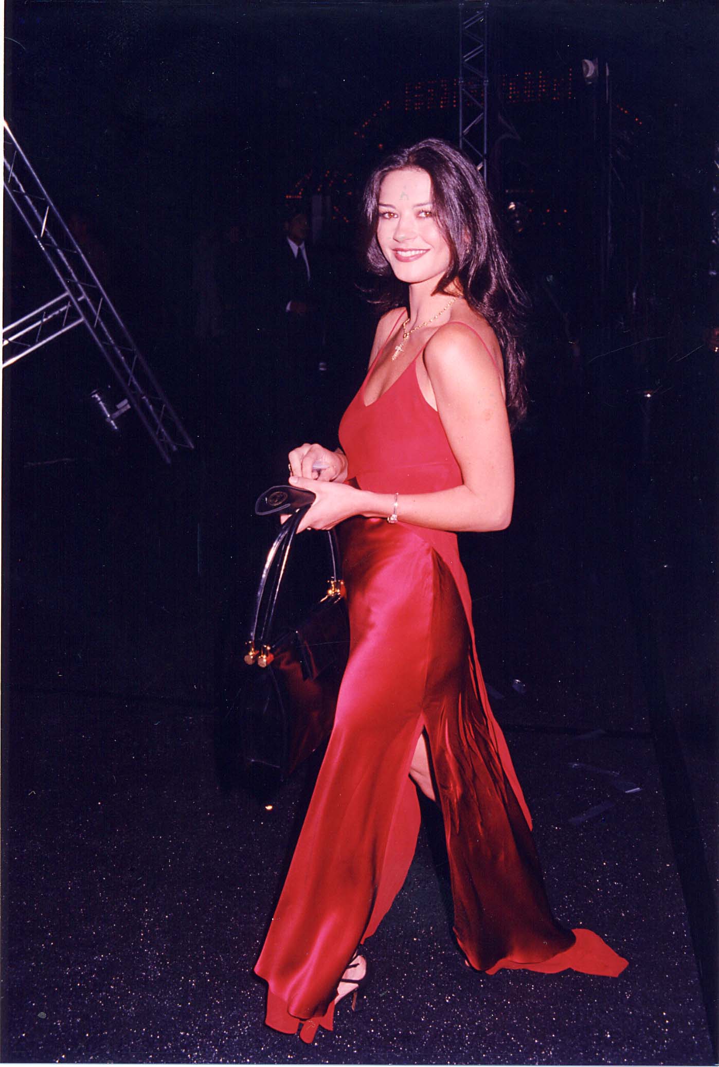 Catherine Zeta-Jones turns with a smile during the premiere of "Men in Black" on September 6, 1997, in Los Angeles, California. She arrives in a flowing crimson spaghetti-strap gown designed with dual thigh-high side slits, holding a black leather handbag and accessorizing with strappy heeled sandals and a delicate gold cross necklace.