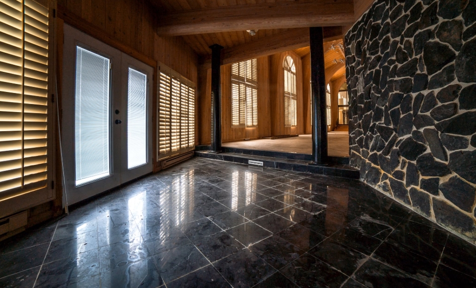 Polished dark flooring leads into a soaring wood-lined room at Mike Tyson's old residence, while a rugged stone wall provides a sharp contrast along one side. Beyond the raised threshold, sunlight filters through tall shuttered windows beneath exposed wooden beams. The mixture of stone, timber, and marble-like finishes showed the varied materials incorporated throughout the heavyweight champion's enormous former home. | Source: Facebook/Johnny Joo