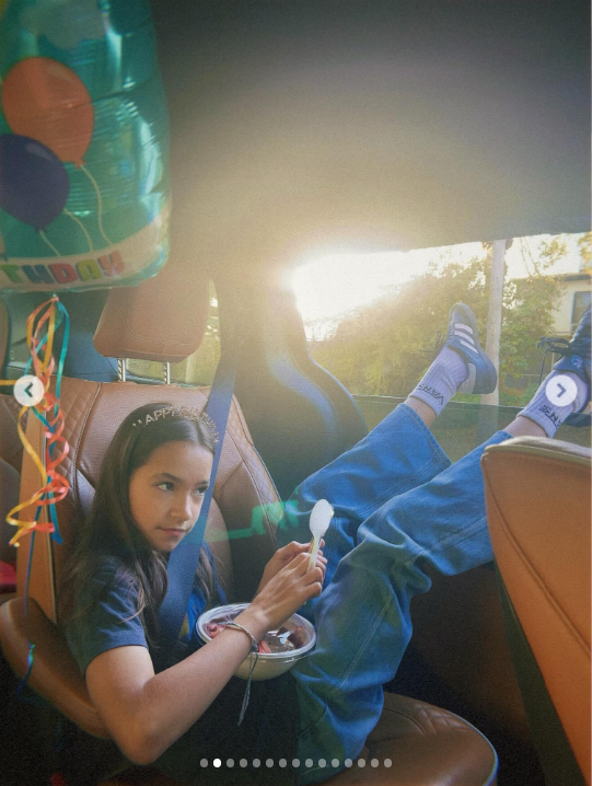 Fiona is pictured alone in the back seat of a car, wearing a dark blue T-shirt and blue jeans with white socks and sneakers. With her legs stretched across the seat, she holds a spoon over a small container of food in her lap, while sunlight streams through the window and colorful balloon decorations hang nearby.