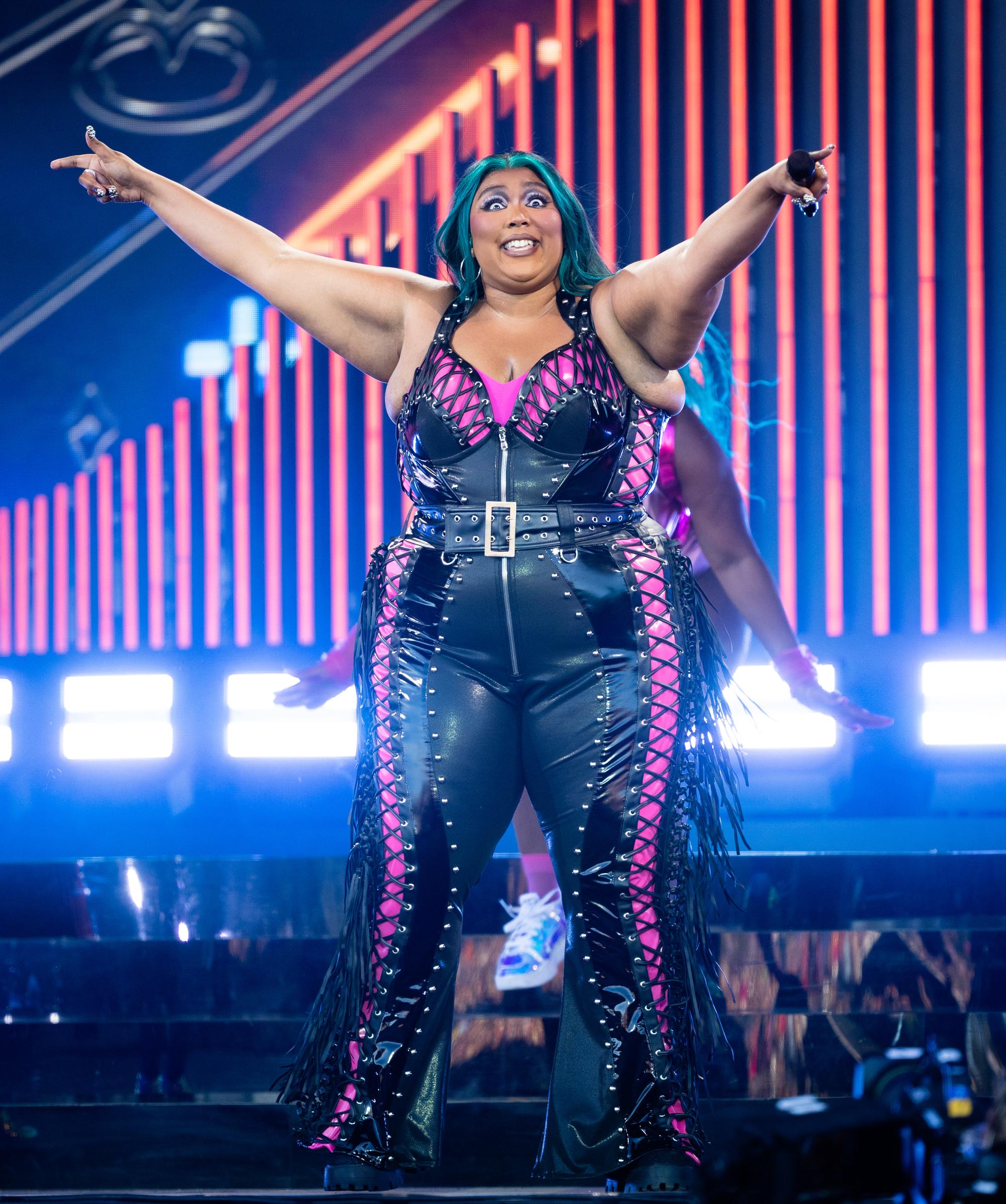 During a performance at Glastonbury, Lizzo wore a black leather-look corset jumpsuit with pink lace-up paneling and fringe details. The belted design cinched her waist while drawing attention to her larger arms, wide hips, and visibly heavier frame.