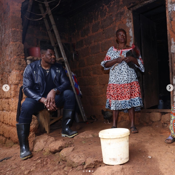 Seated beside the entrance of a weathered brick home, Francis Ngannou looked off to the side while a woman stood nearby and a white bucket rested in the foreground. The scene offered a glimpse of the surroundings in Batié, where his early years had been marked by poverty and physically demanding work before he eventually left Cameroon in pursuit of a professional fighting career. | Source: Instagram/francisngannou