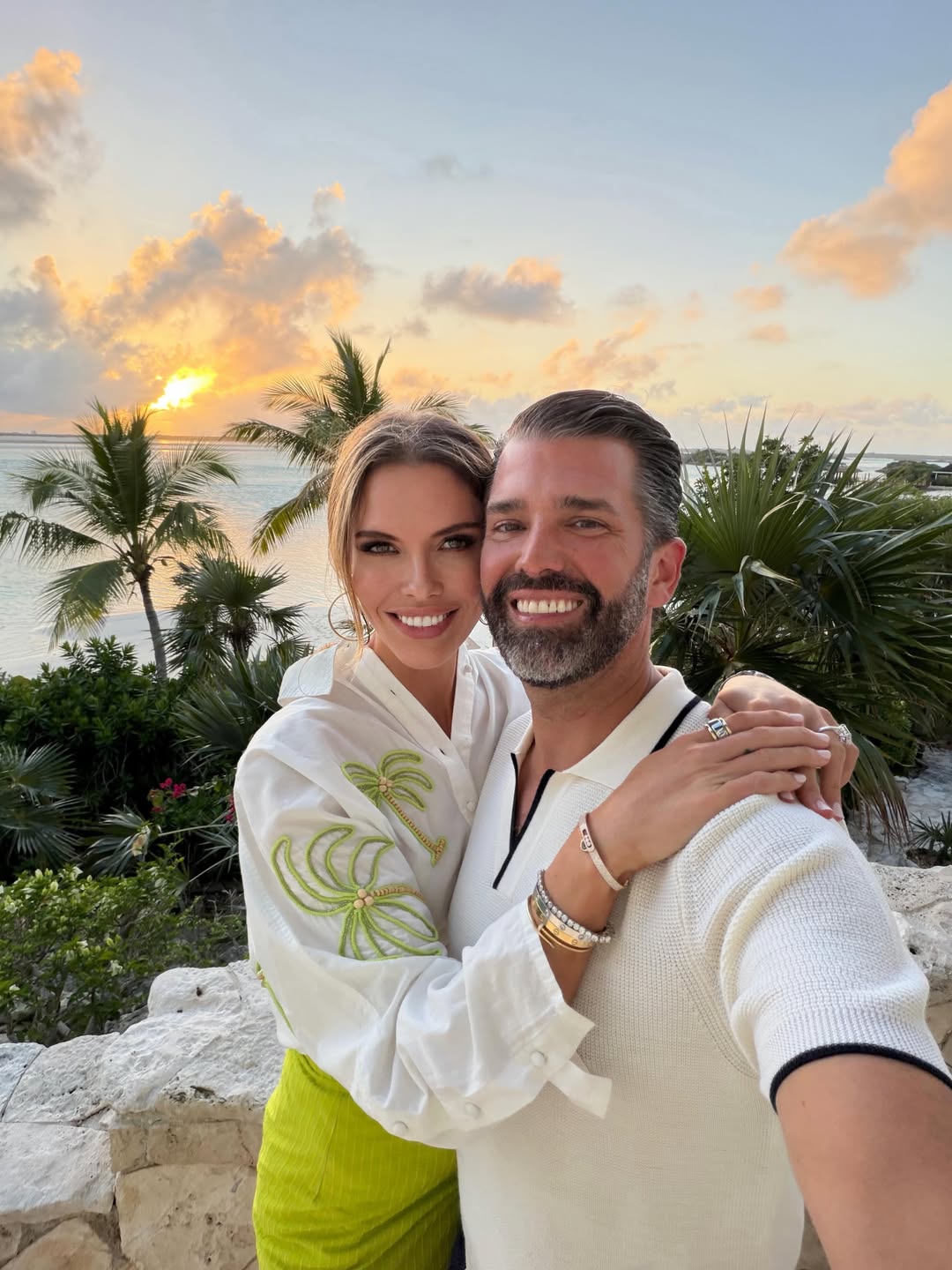 Donald Trump Jr. and Bettina Anderson pose arm in arm at sunset, with golden light reflecting across the ocean behind them.