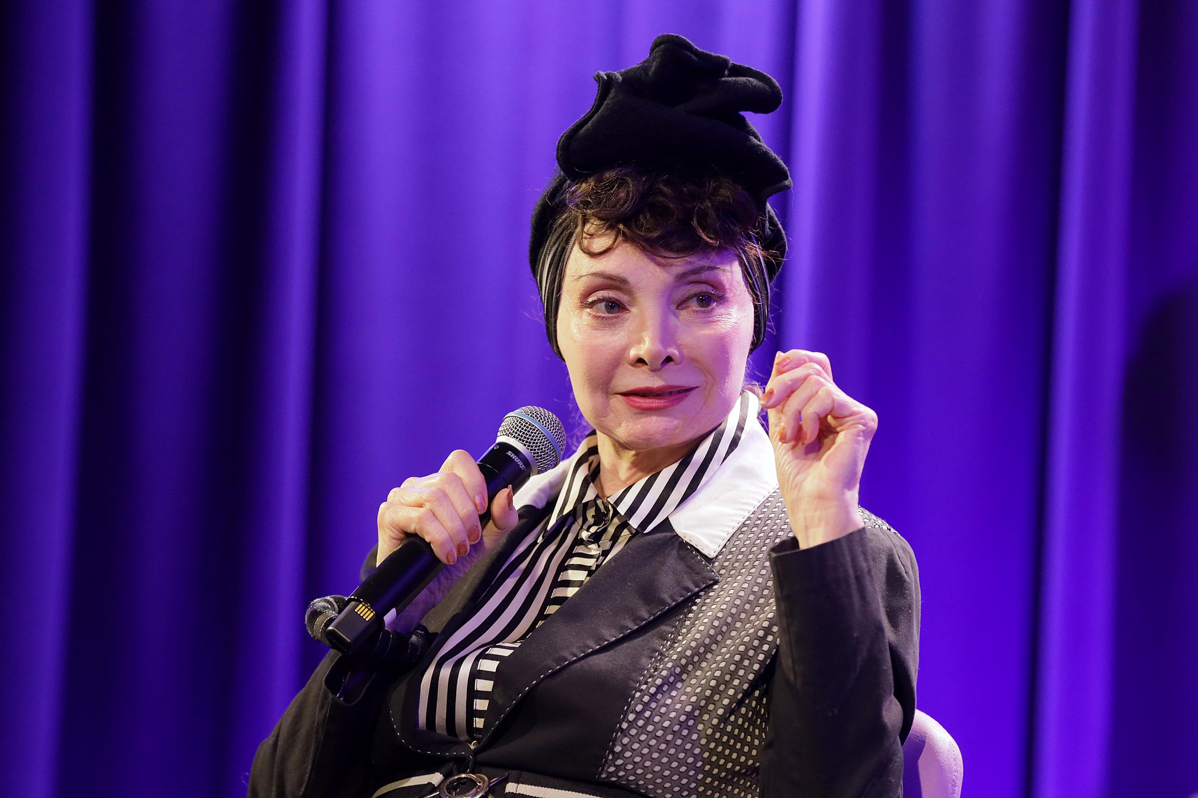 Toni Basil speaks into a microphone during a discussion celebrating the 40th anniversary of her hit song “Hey Mickey” at the GRAMMY Museum in Los Angeles, wearing a black-and-white outfit and a distinctive black headpiece. | Source: Getty Images