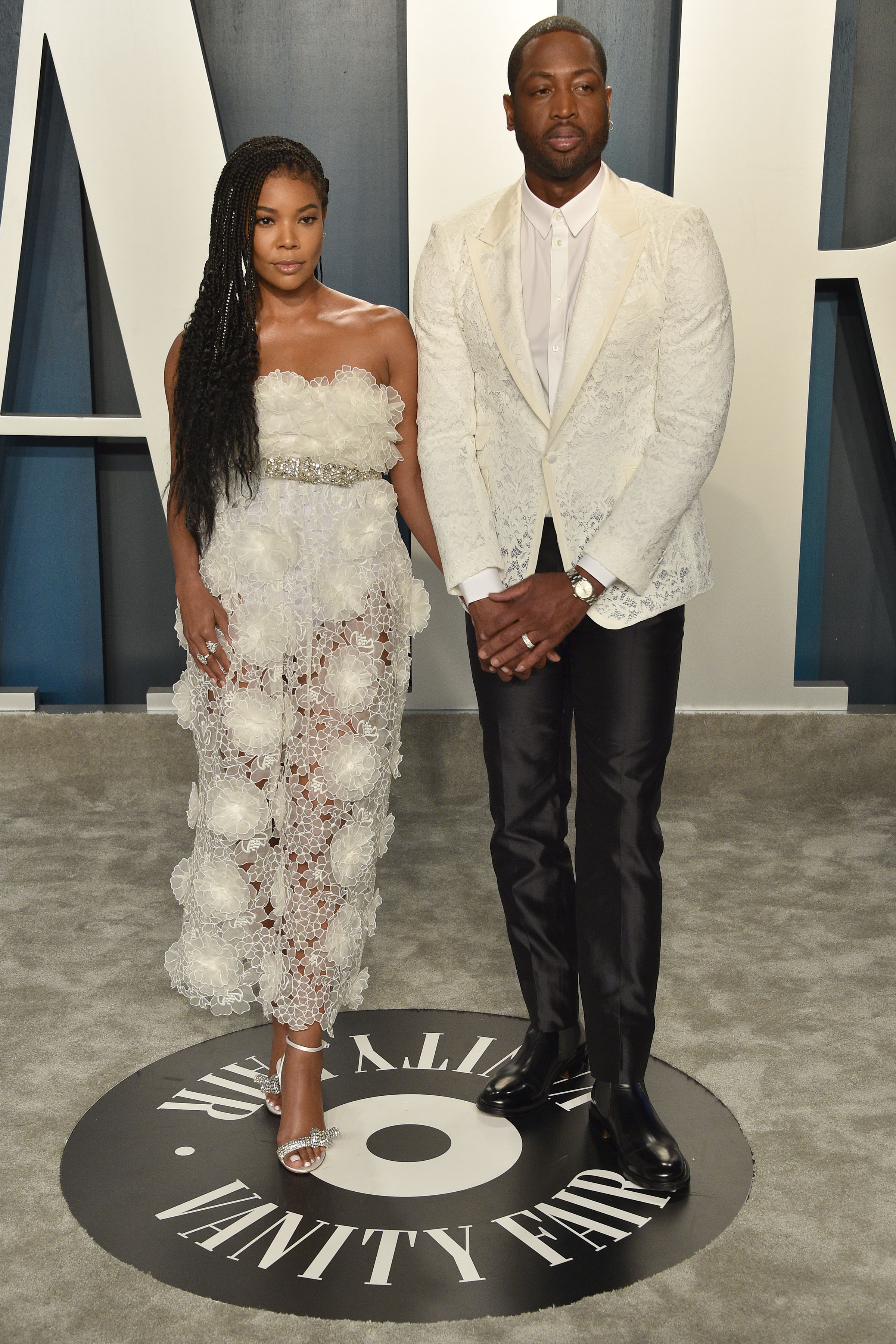 Gabrielle Union stands alongside Dwyane Wade at a formal event. She wears a white, intricately detailed strapless dress with floral textures, while he’s dressed in a patterned white jacket and black trousers. | Source: Getty Images