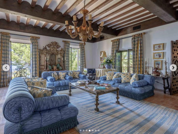 Exposed ceiling beams, patterned curtains, antique-style furnishings, and blue upholstered sofas give this living room a layered, traditional look. Large windows on multiple sides keep the space connected to the lush landscape beyond the walls. | Source: Instagram/robbreport