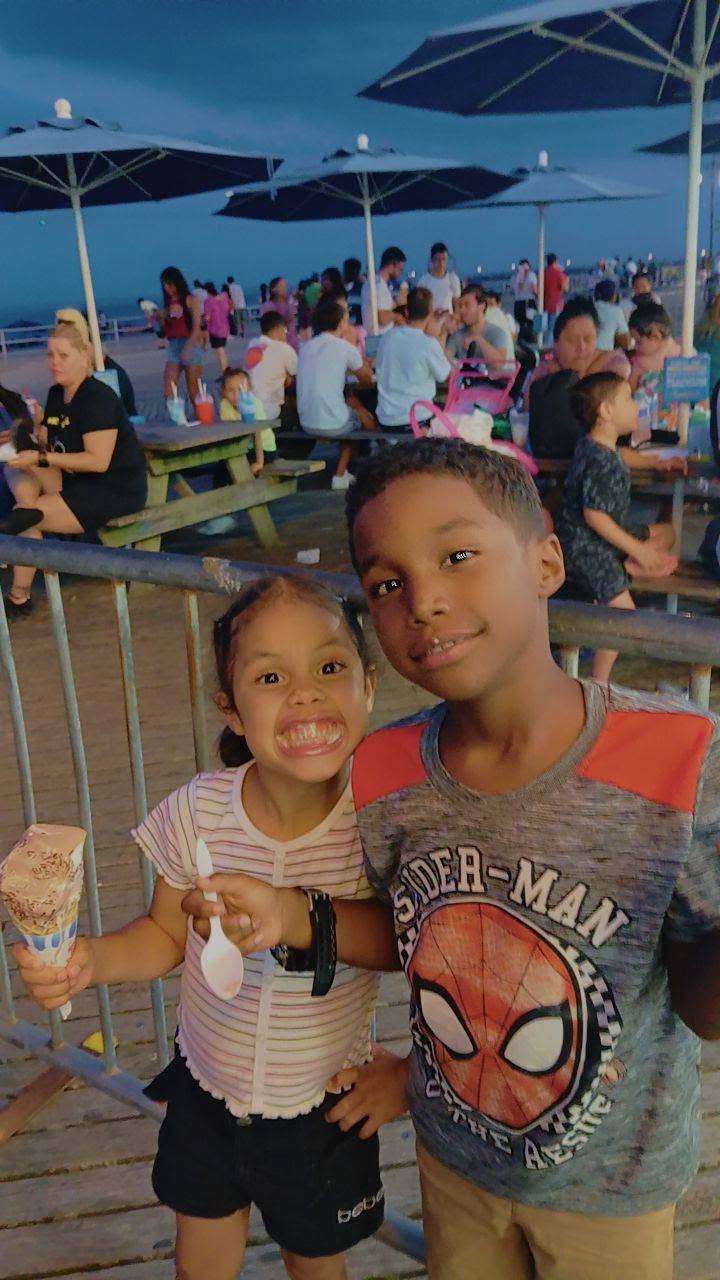 Eddimar Paola Hernández Chacón showed off a big grin while enjoying an ice cream cone alongside her brother at an outdoor boardwalk patio. She wore a striped short-sleeve top paired with black shorts, posing beside her brother, who sported a grey "Spider-Man" graphic t-shirt against a lively backdrop of evening diners under patio umbrellas. | Source: Facebook/Morena Esa