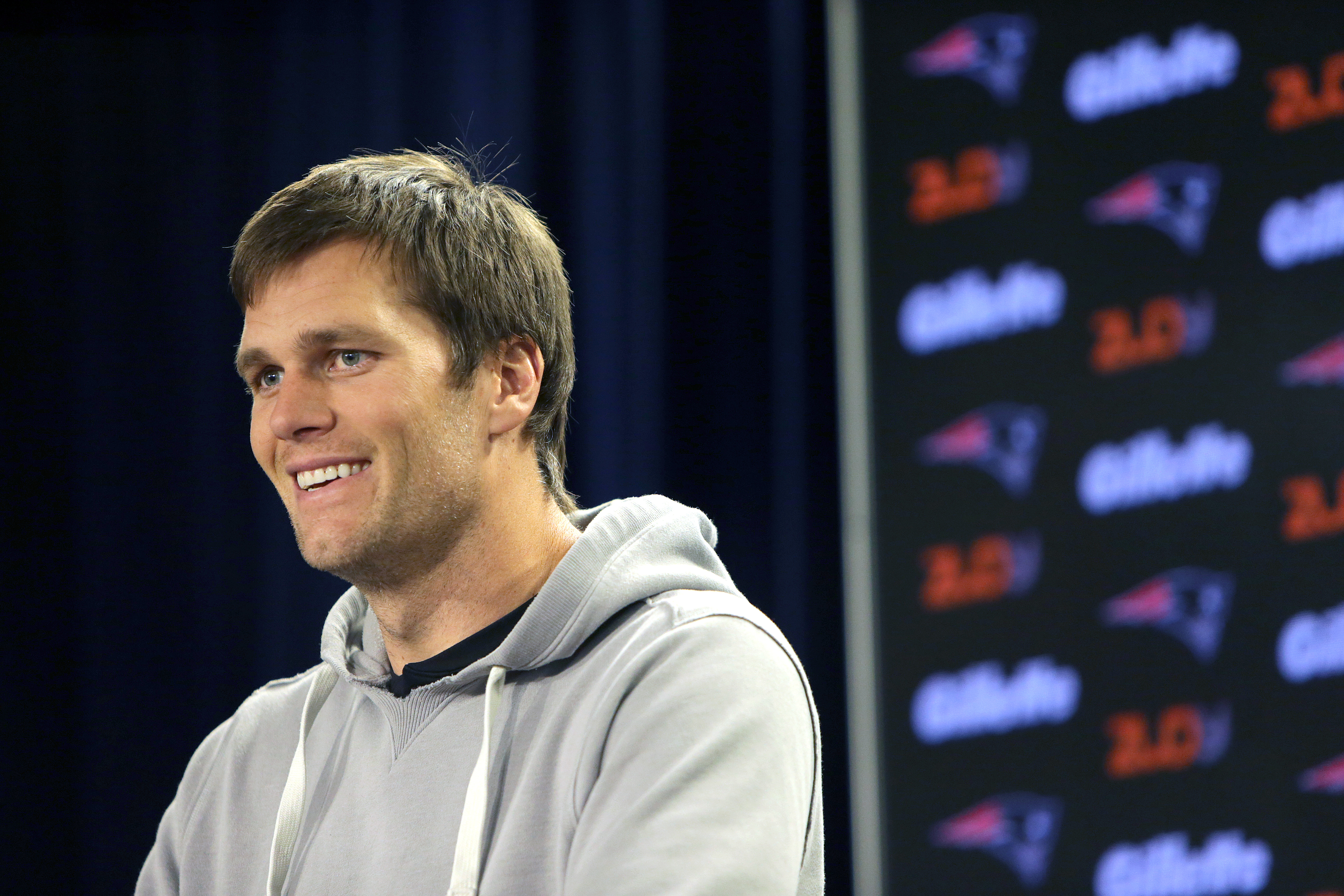 Tom Brady, sporting a gray hoodie over a black T-shirt, shares a light moment at the podium during a New England Patriots practice in Foxborough, Massachusetts. Caught mid-laugh, he looks relaxed and charismatic as he engages with the media.
