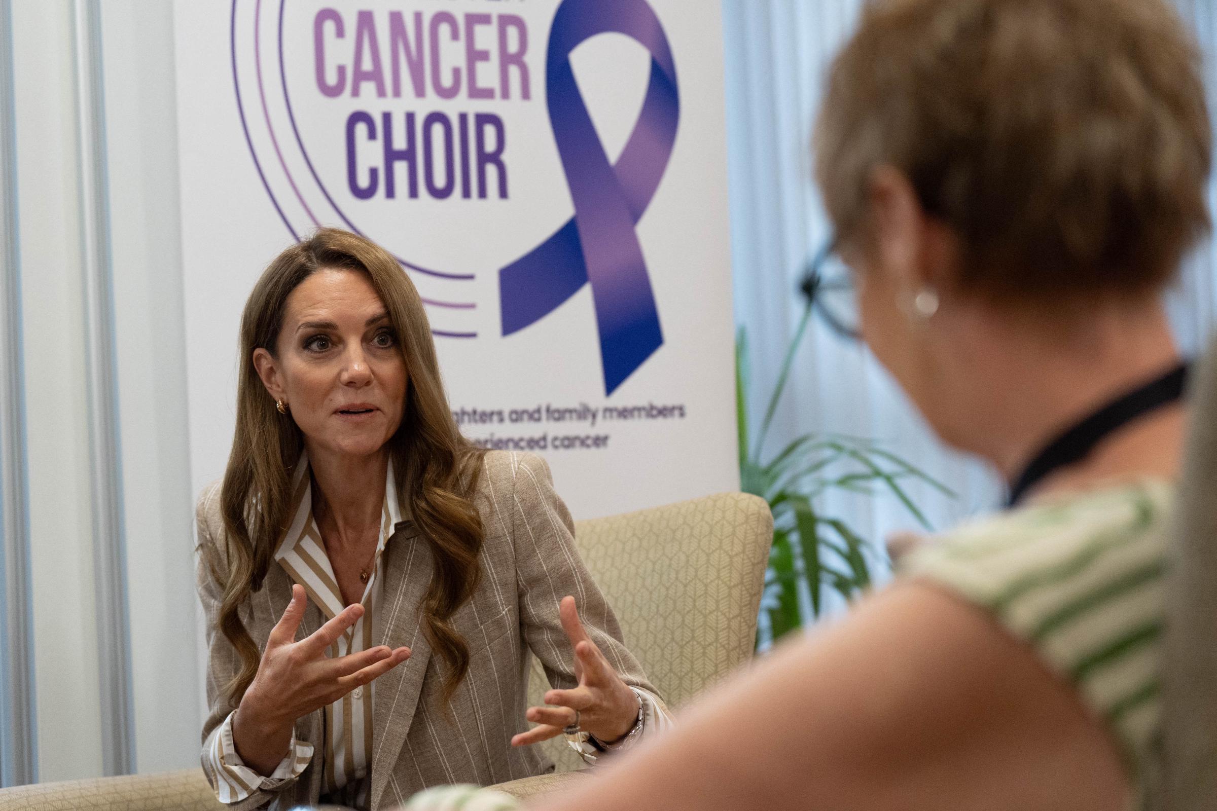 Seated beside a cancer awareness sign, Princess Catherine speaks with a guest. The Princess of Wales's expression is engaging and sincere.