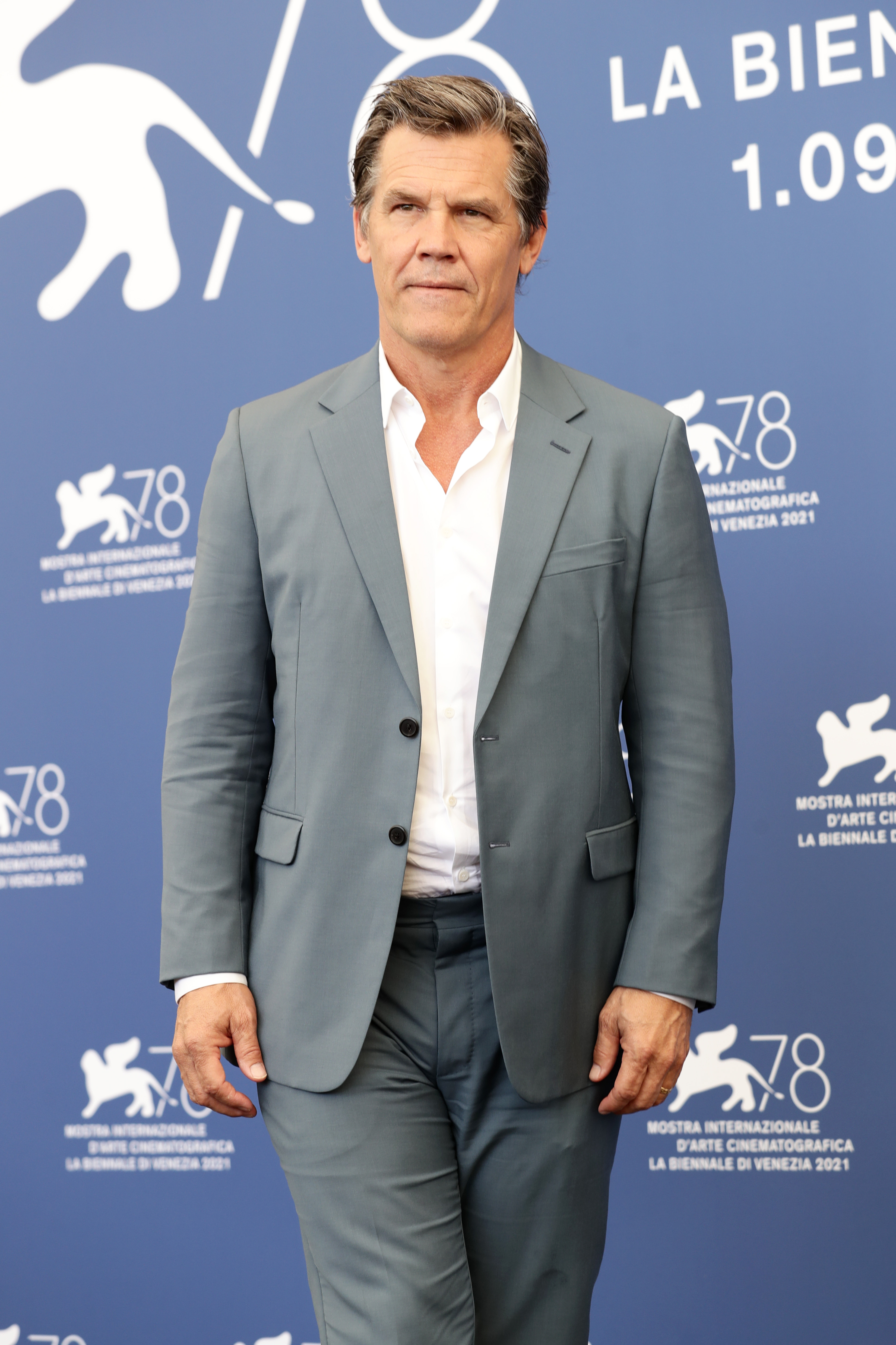 Wearing a white dress shirt and navy tie beneath a dark suit, he appears slightly aged but clean-shaven, with a short, natural haircut and visible signs of graying, a reserved, formal look with no tattoos visible.