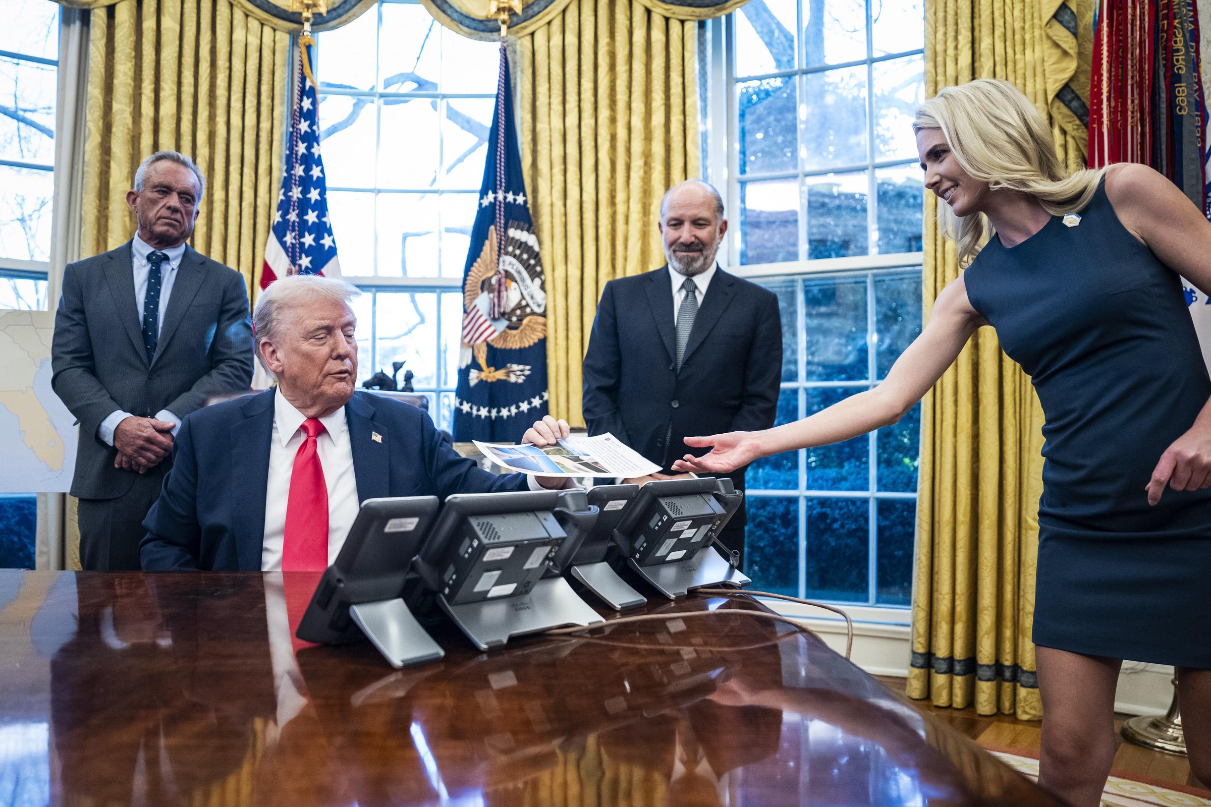 Donald Trump sits at the Resolute Desk as aide Natalie Harp hands him documents during a 2025 White House meeting. Her role as the aide who kept information flowing directly to the president became central to her unusual proximity — and eventually helped fuel the nickname colleagues gave her. | Source: Getty Images