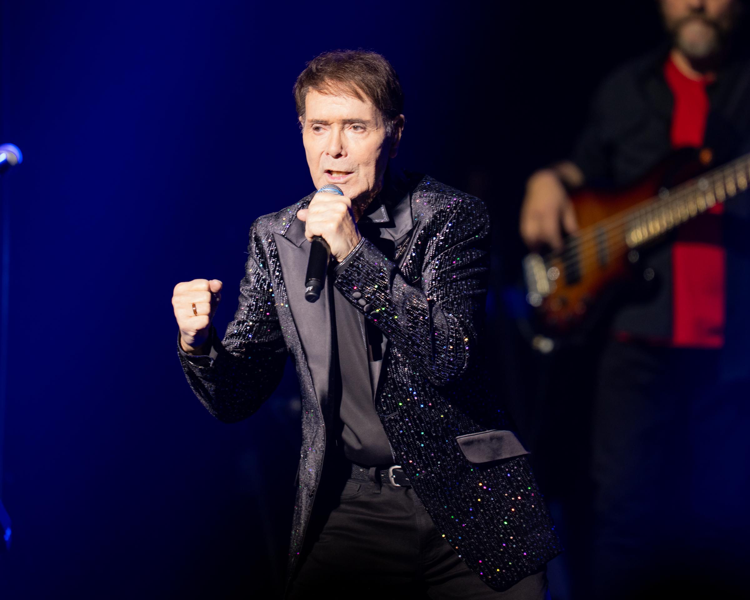 Cliff Richard performs under vivid blue stage lights, years after his friendship with Bonnie Tyler and Robert Sullivan extended beyond Portugal to holidays and dinners together in Barbados. | Source: Getty Images