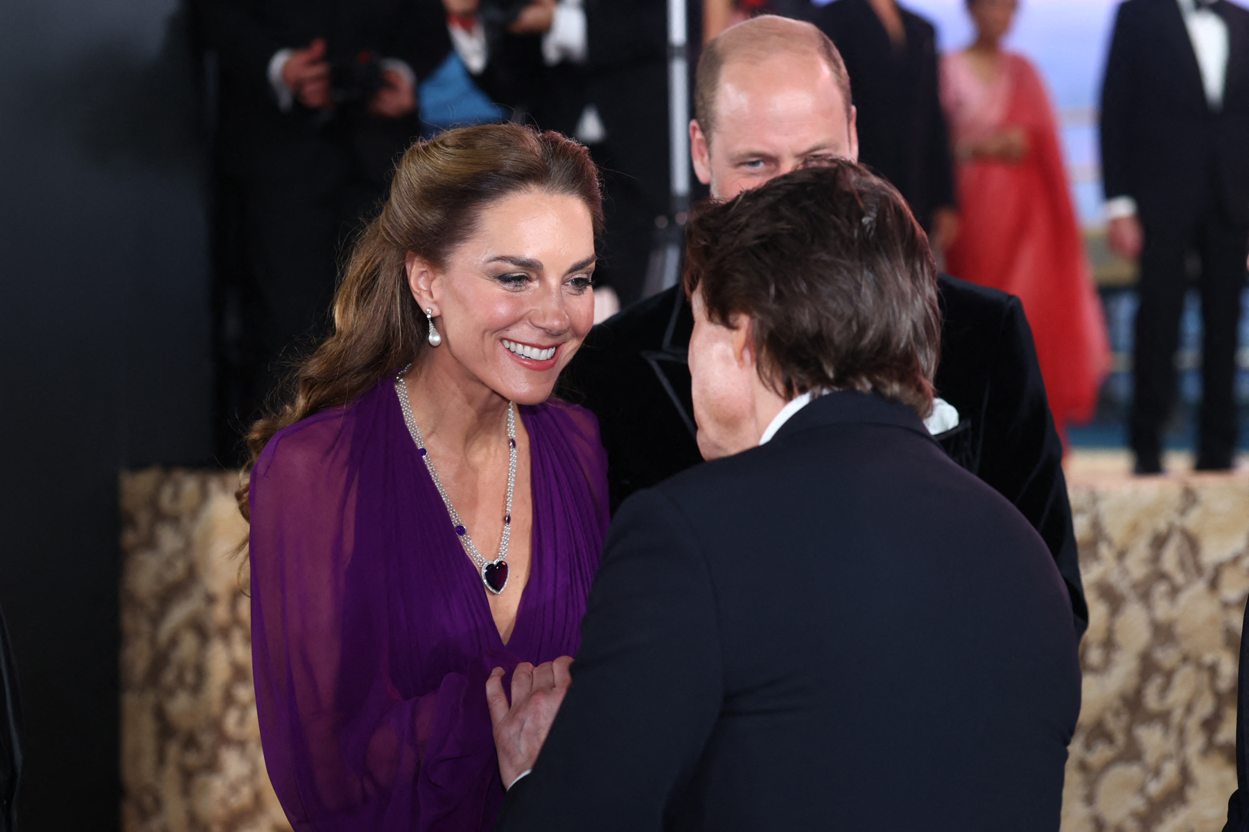 Catherine speaks with Tom Cruise as William stands behind them at the "Digger" global premiere in Leicester Square. Her dark pendant necklace and pearl drop earrings are visible against the plunging neckline of her purple gown.
