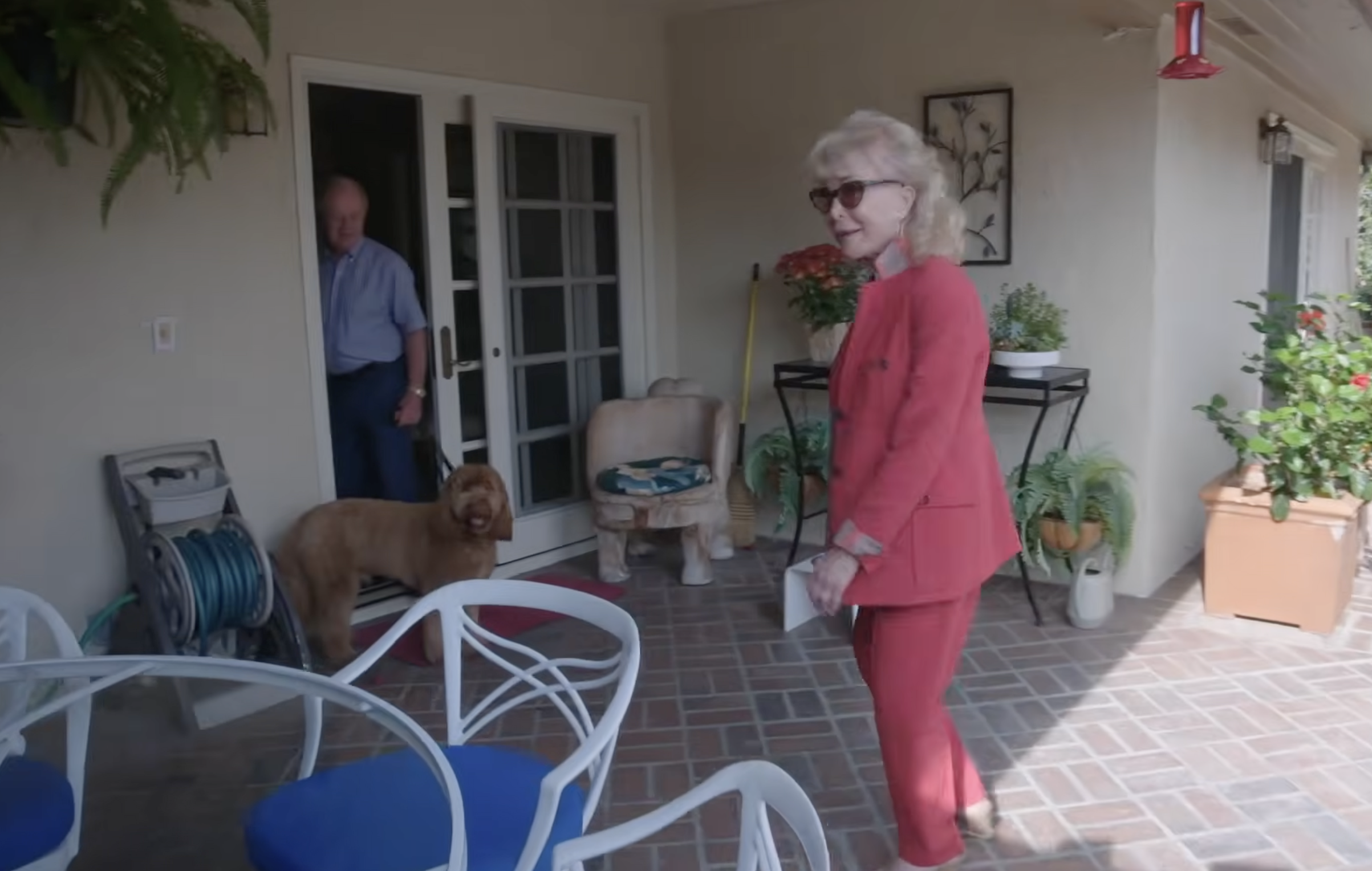 Barbara Eden turns toward the camera on her patio, wearing oversized sunglasses and her bright pink-red suit. Her husband stands in the doorway behind her as their fluffy brown dog waits nearby, surrounded by potted plants and outdoor furniture. | Source: YouTube/People