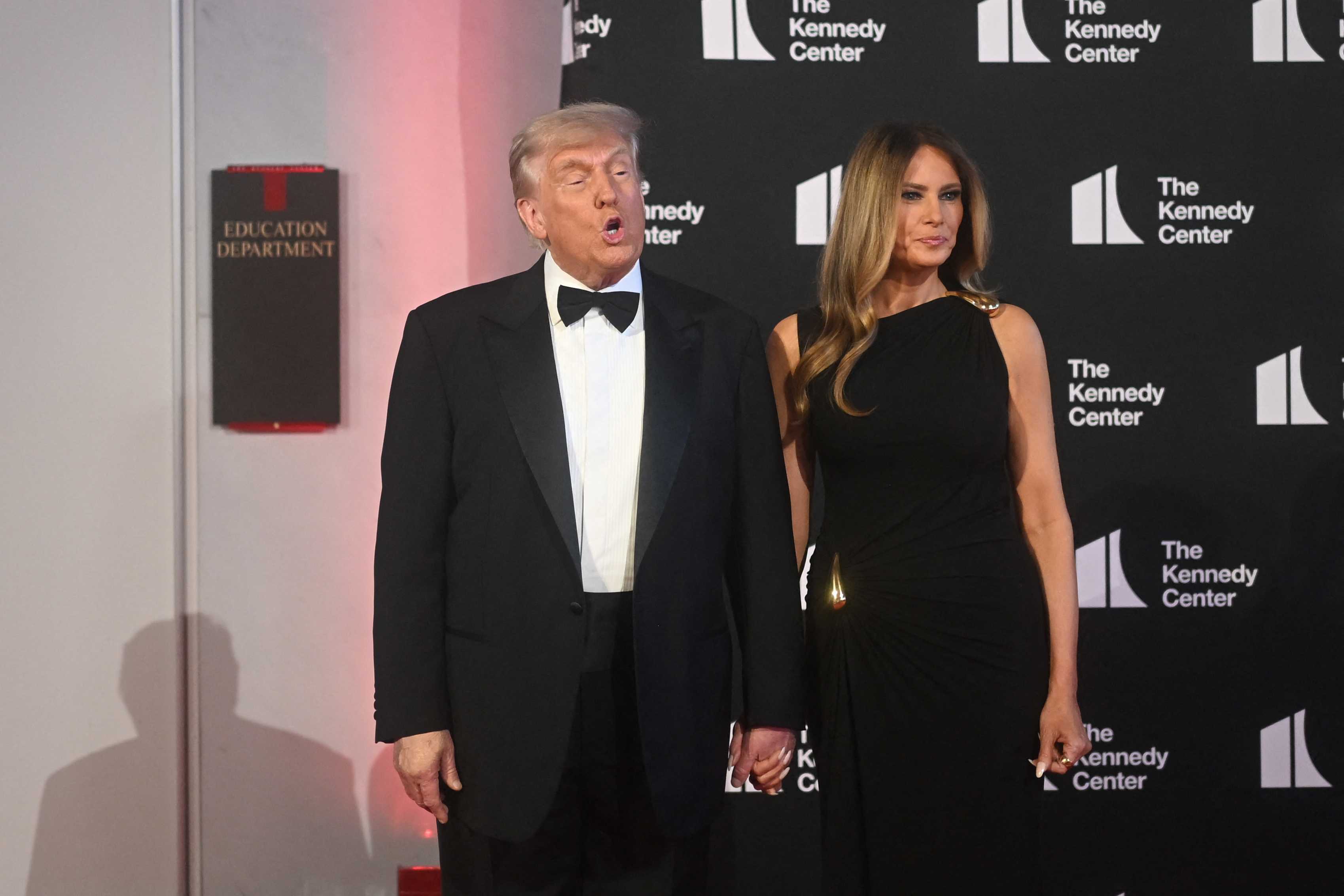 Standing beneath a sign for the Education Department, the president appears mid-sentence while the first lady maintains a composed look as they pose together at the Kennedy Center on June 11, 2025.