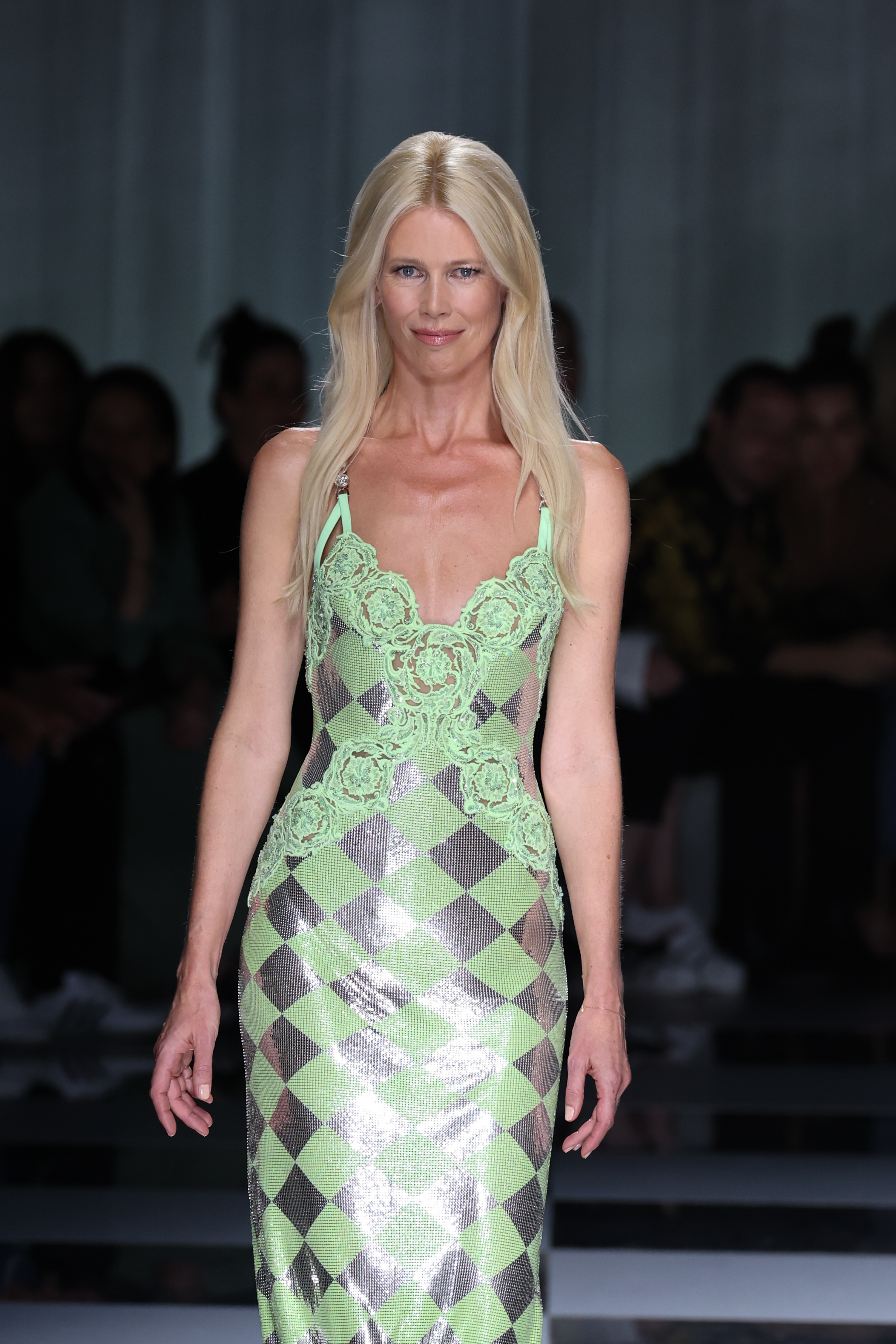 Claudia Schiffer makes a dazzling return to the runway at Versace's Spring/Summer show in Milan, glowing in a metallic green-and-silver checkered gown adorned with lace detailing. Her confident stride and timeless beauty electrify the fashion week spotlight.