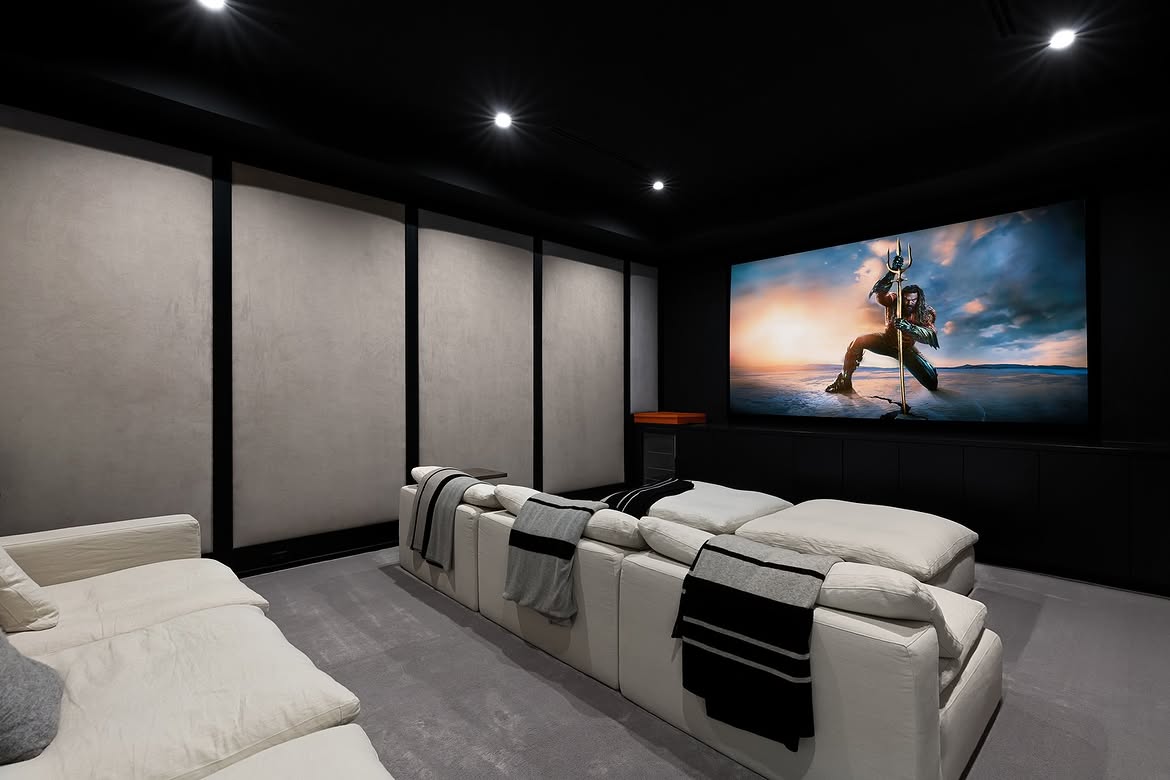 The home's private theater shifts away from the bright palette used throughout the main living spaces. Black walls and ceilings surround rows of oversized white seating, while recessed lighting keeps the focus on the large screen. | Source: Instagram/tomer.fridman