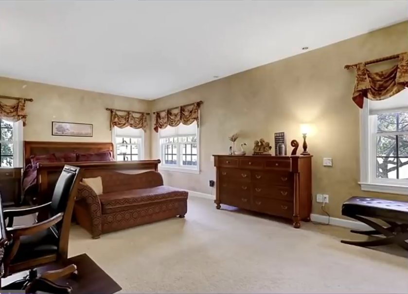 The bedroom features beige walls, cream carpeting, and dark wooden furniture, including a bed, a matching dresser, and upholstered seating. Ornate patterned window valances and heavy furnishings give the room a formal, traditional appearance.