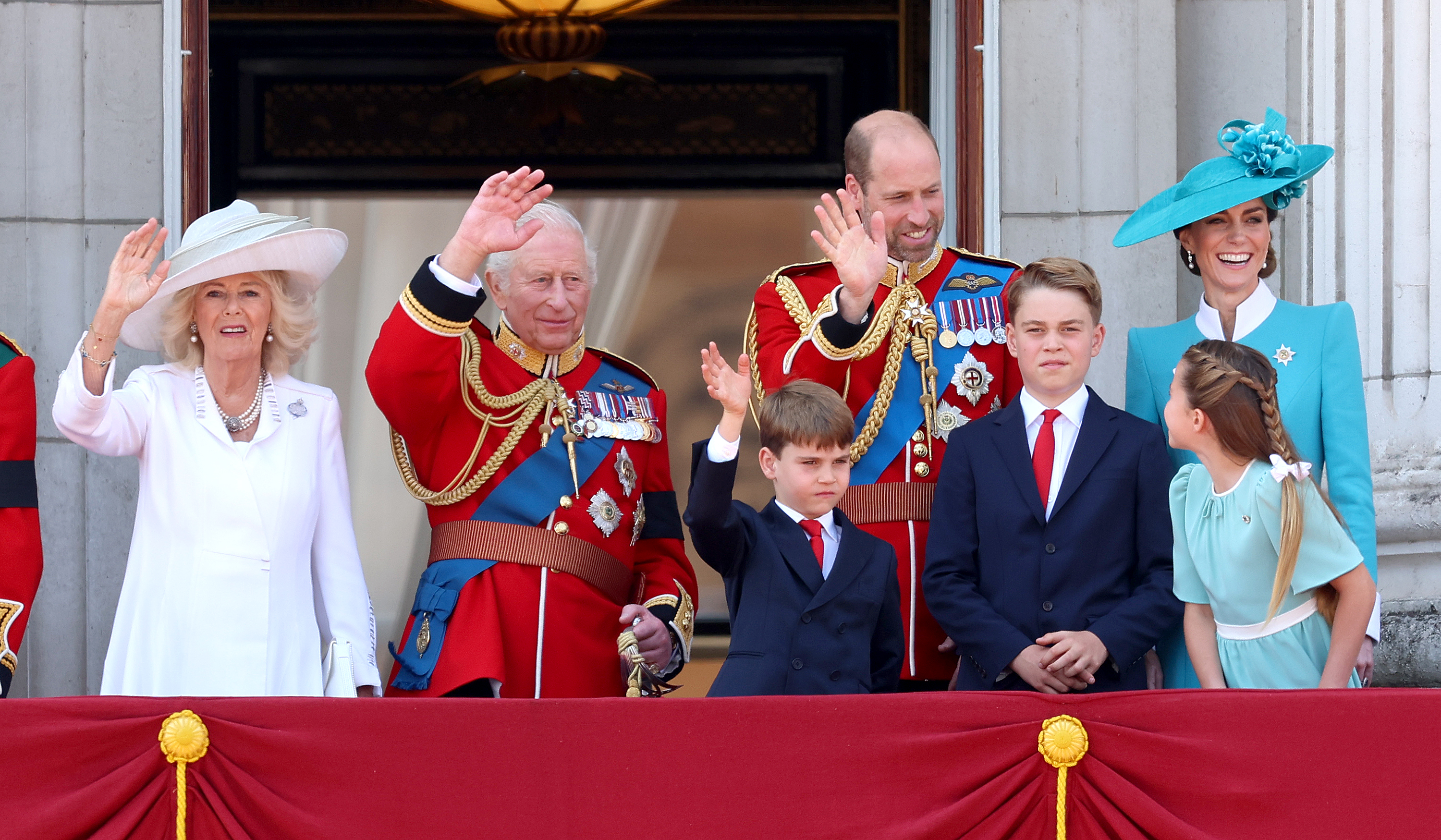 Queen Camilla, King Charles III, Prince Louis, Prince William, Prince George, Princess Charlotte, and Princess Catherine greet the crowd during the 2025 Trooping the Colour ceremony. Princess Charlotte looks back at her father, Prince George poses with his wrists crossed, Princess Catherine smiles, and the other royal family members wave to the crowd.
