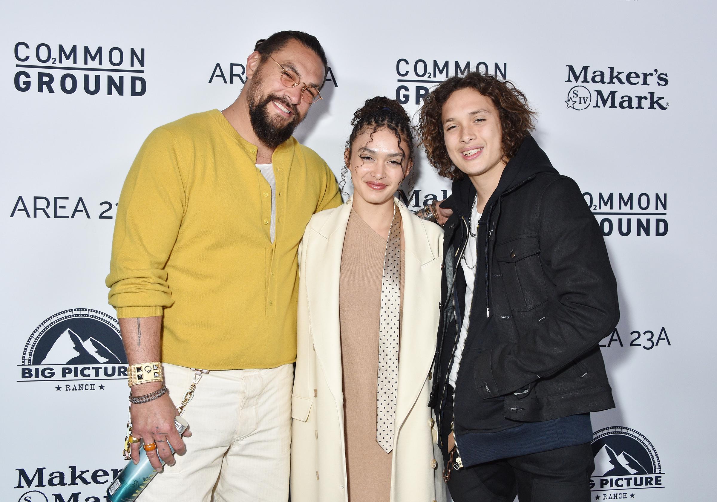 Jason Momoa poses alongside his children Lola Iolani Momoa and Nakoa-Wolf Manakauapo Namakaeha Momoa at the Los Angeles special screening of "Common Ground" at the Samuel Goldwyn Theater on January 11, 2024, in Beverly Hills, California. Momoa sports clear-framed glasses, a bright mustard-yellow knit henley, and cream trousers while holding an aluminum water bottle, standing next to Lola in a beige dress with an ivory coat and dotted scarf, and Nakoa-Wolf smiling in a dark hooded jacket layered over a denim trucker jacket. | Source: Getty Images