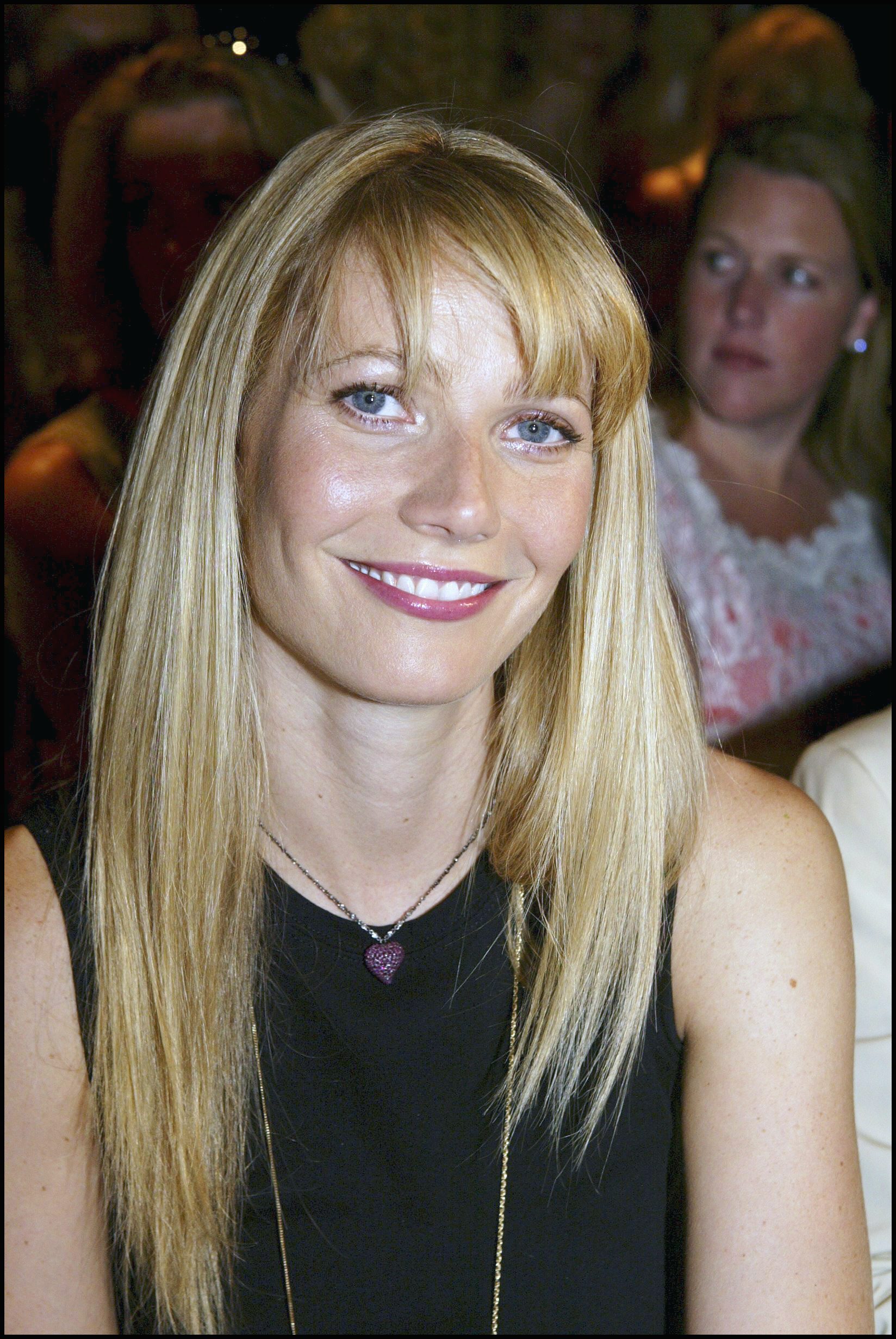 Gwyneth Paltrow attends the Valentino Haute Couture Fall-Winter 2003-2004 show in France on July 9, 2003. She wears a sleeveless black top paired with a delicate purple heart pendant necklace. Her straight blonde hair is styled with soft bangs that frame her face, while her radiant smile and glossy berry lip add a polished touch to her look.
